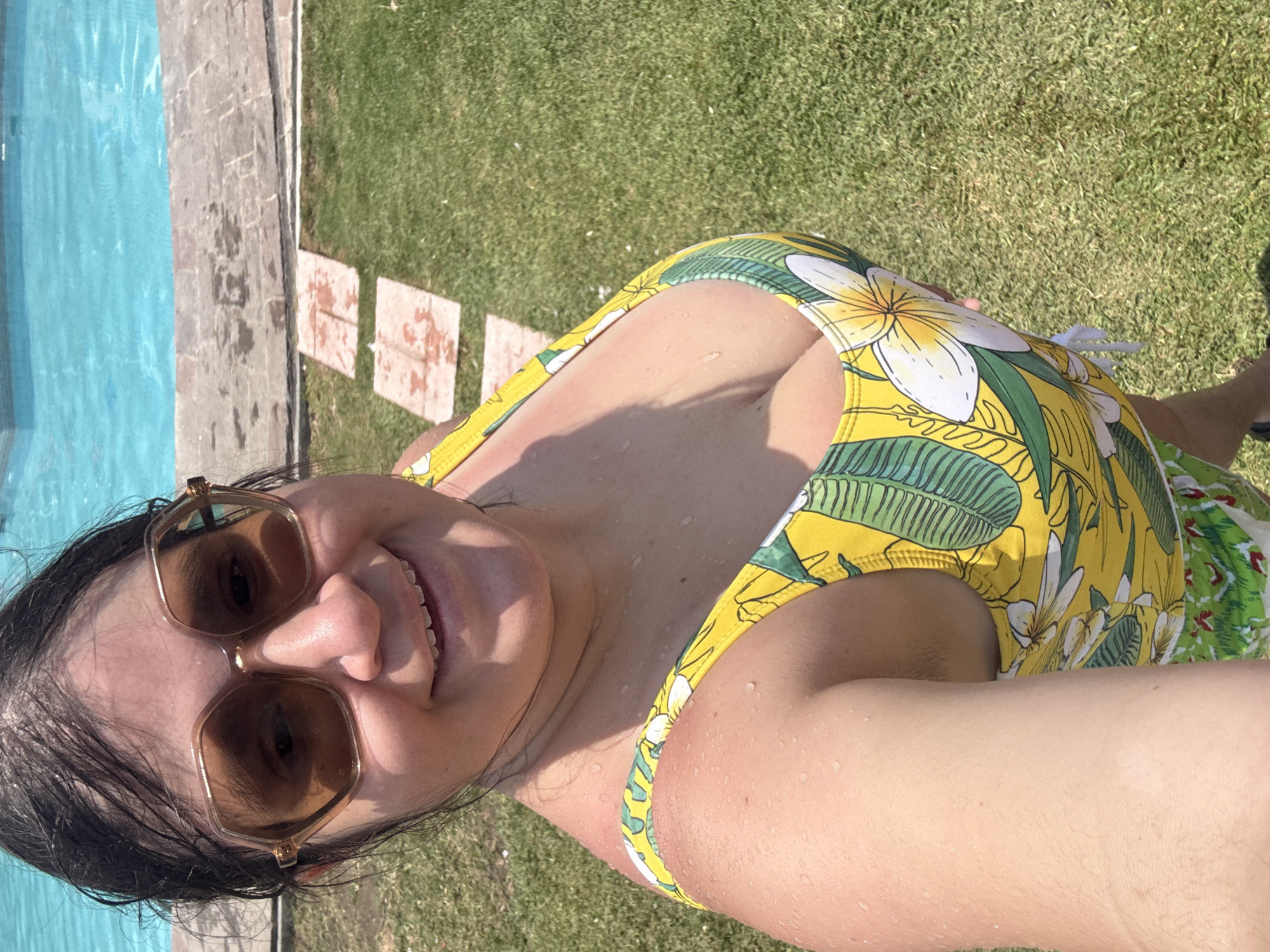 A woman captures a selfie outdoors on a bright, sunny day, likely enjoying leisure time by a swimming pool in Tres de Mayo, Mexico. She is positioned in the foreground, smiling broadly at the camera. Her dark hair appears slightly wet, and water droplets are visible on her skin and shoulders, suggesting she has recently been in the pool or splashed. She wears stylish brown-tinted sunglasses with angular frames.
Her attire is a vibrant yellow swimsuit adorned with a tropical pattern of large green leaves and white frangipani-like flowers. The swimsuit features a plunging neckline.
The background reveals a clear blue swimming pool to her left, with a paved or stone path separating the pool from a patch of lush green grass. The path shows some darker, possibly wet, spots. The overall scene suggests a warm, clear day, with bright sunlight creating visible shadows. No text is visible in the image.