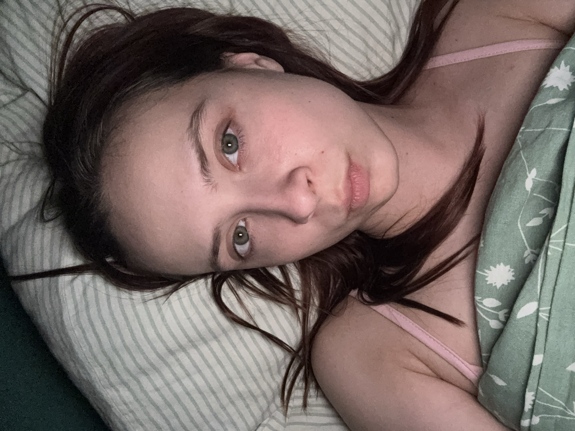 A woman lies on a striped pillow with a green and white floral-patterned blanket pulled up to her chest. She has fair skin, green eyes, and brown hair. She wears a pink tank top. The location is unknown; there is no evidence the picture was taken in Прикамский, Russia.