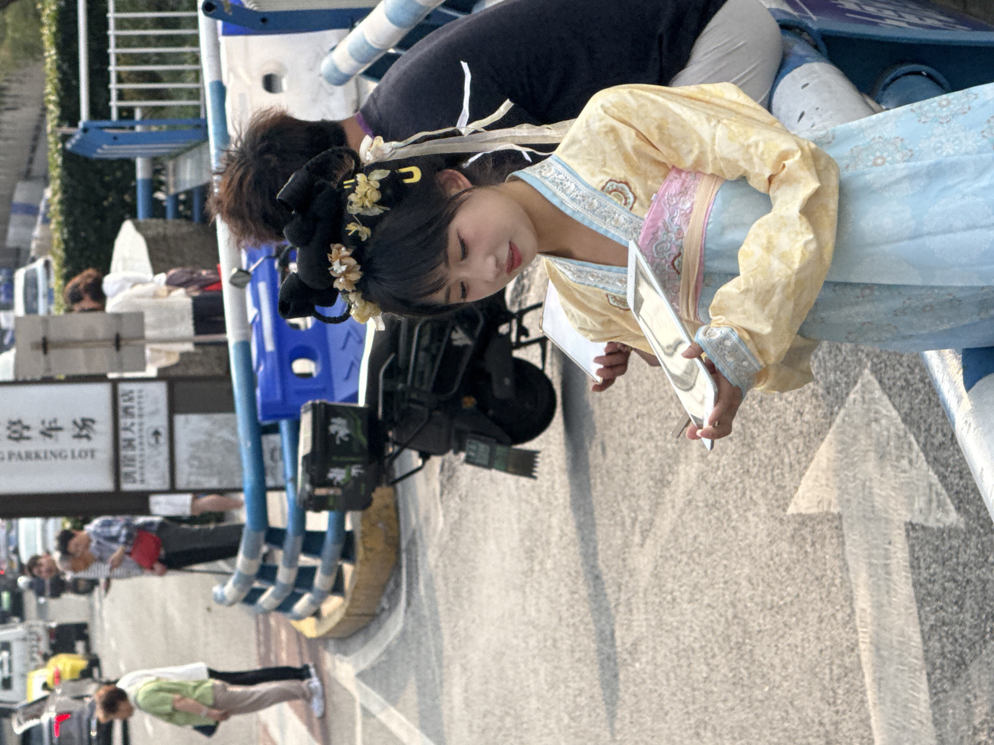 In the Yuzhong District of China, a woman in traditional Hanfu attire, with a yellow and blue garment, is holding a small, rectangular mirror. She is standing on a paved road marked with a white painted arrow. Behind her, a man with dark hair is partially visible, and a professional film camera mounted on a rig. Further in the background are other pedestrians, parked vehicles, and a sign displaying Chinese characters and the English words "PARKING LOT". The overall setting suggests an urban environment with a filming activity taking place.