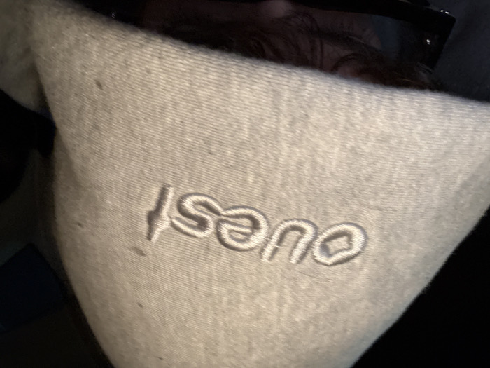 A close-up image taken in Givors, France, shows an individual wearing a light grey hooded garment. The word "ouest" is embroidered in white thread on the upper right side of the hood. The hood covers the upper portion of the person's face, with dark hair and dark-rimmed sunglasses visible below its edge. The immediate background is dark and indistinct, suggesting a low-light or indoor setting. No specific actions or interactions are discernible beyond the individual wearing the apparel.