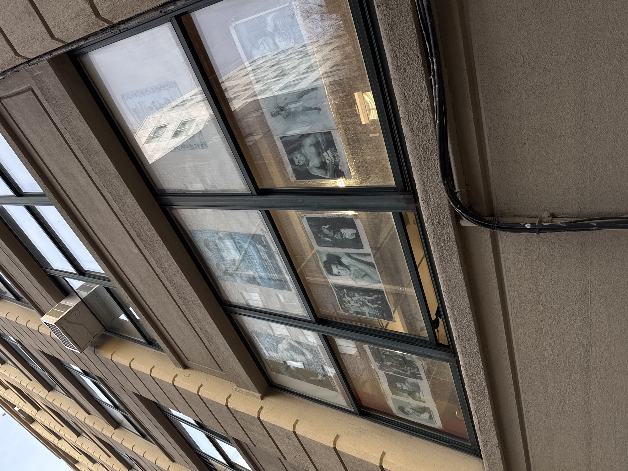 The image depicts the windows of a building, angled upwards, with posters or images visible behind the glass. The building is constructed of a light brown material with a textured surface and a repeating rectangular pattern along its facade. The windows are framed in black. A reflection of another building is visible in one of the windows. A black cable or conduit runs along the building's exterior. From information known from other sources, this is the windows of Gleason's Gym in DUMBO, New York City, known for its boxing history and posters.