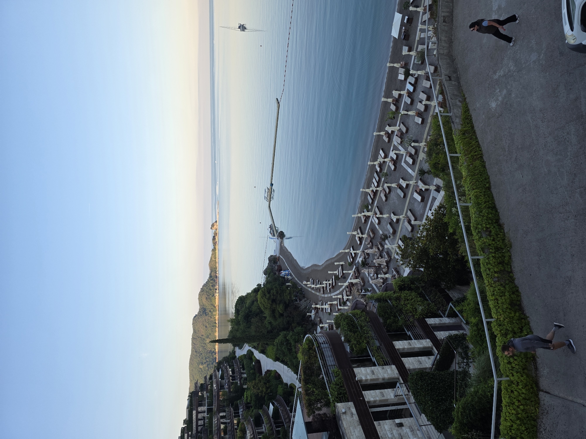 The image captures a panoramic view of a resort area on the coast of Budva, Montenegro. In the foreground, a paved walkway slopes downwards, with a man in athletic wear jogging on the left and a woman walking on the right. Both are seen from a high vantage point.
Dominating the middle ground is a terraced resort building, characterized by its beige stone facade and dark-framed windows, draped in lush green foliage. Adjacent to the building is a well-maintained beach with rows of empty lounge chairs and umbrellas, facing a calm, blue sea. A wooden pier extends into the water, with a small boat moored beside it.
Further out in the sea, a sailboat is visible on the horizon. The background features a chain of tree-covered hills and mountains, with the coastline curving into the distance. The sky is clear and a pale blue, with a hint of lighter hues near the horizon, suggesting early morning or late evening light. The overall scene depicts a serene and upscale coastal vacation setting.