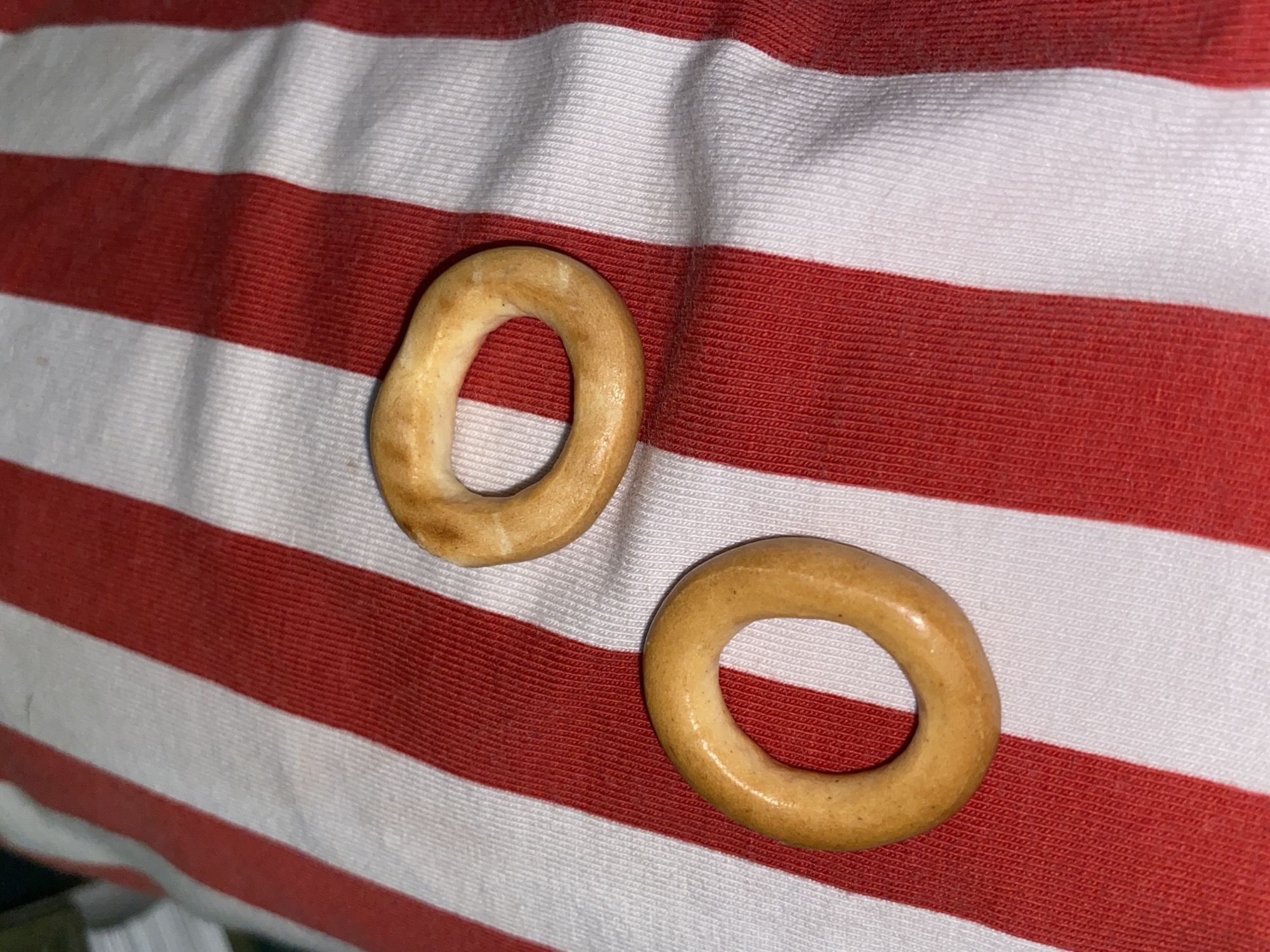 The scene, located in the city of Chaikovsky, Russia, displays two ring-shaped baked bread products, consistent with sushki or baranki, positioned on a textile surface. The background fabric is characterized by distinct alternating vertical stripes of red and white. The upper-right bread ring is light golden-brown, exhibiting an slightly irregular, somewhat oval shape with a minor indentation on its top curve. The lower-left bread ring is also light golden-brown, smaller, and more uniformly circular. Both items feature a smooth, lightly reflective surface. The fabric exhibits a visible woven texture and minor creasing. No human subjects or actions are depicted.