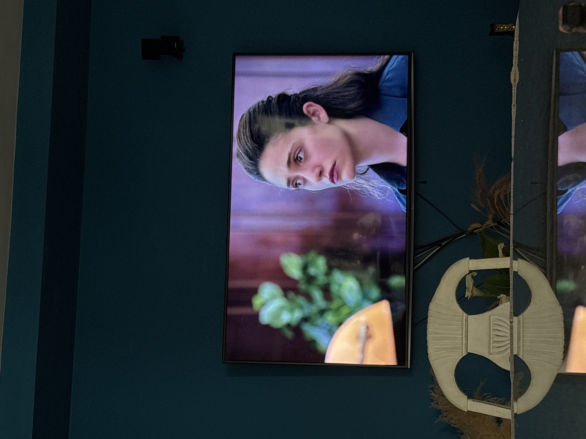 An indoor scene features a television screen mounted on a dark teal or blue wall. The TV displays a close-up of a young woman with dark hair, styled partially pulled back. She wears a dark blue garment and has a serious or contemplative expression, looking downwards and to her left. Her face is pale, and her gaze appears intense. In the background of the television screen, blurry green foliage and a yellow object (possibly a piece of food or decorative item) are visible in the lower right corner, set against a reddish-purple backdrop.
Above the television, a small, dark rectangular object, possibly a speaker or camera, is attached to the wall. To the right of the television, a large, ornate white decorative object or mirror hangs on the wall, reflecting parts of the room or the TV screen. This object has a distinctive, almost geometric, segmented design with central horizontal ridges and circular openings. Dried, feathery plants or reeds are placed behind and around this decorative piece, adding a natural element to the decor. The overall lighting suggests an indoor setting, likely in low light conditions, possibly in a living area or entertainment space within a residence in Urbanización Encinares De Alcolea, Spain. There is no visible text.
