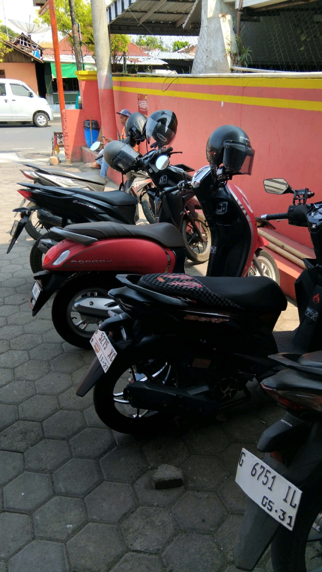 In Batang, Indonesia, a line of parked motorcycles is visible along a reddish-pink wall with yellow horizontal stripes. A white car is parked in the background, and a person wearing a blue cap and jeans is seated on a step near the wall, looking at their phone. Several motorcycles are in the foreground and middle ground, with helmets resting on their handlebars. The ground is paved with hexagonal gray stones. One motorcycle in the foreground has a license plate reading "5275 XC" and "11-27". Another motorcycle in the lower right corner has a license plate reading "G 6751 IL" and "05.31".