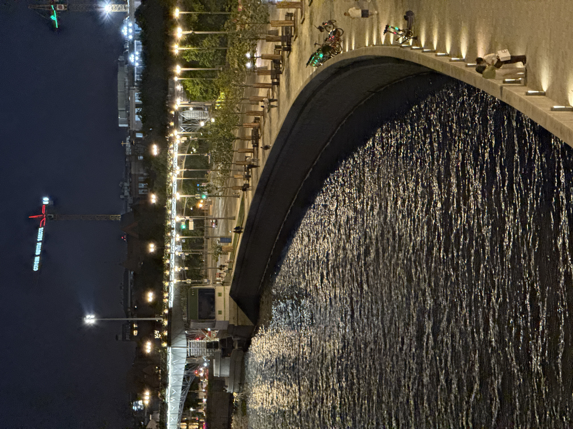 This detailed image captures a night scene along the Moskva River in Moscow, Russia, likely viewed from one of the embankments towards the Zaryadye Park area or a similar central location undergoing development. The scene is illuminated by numerous artificial light sources.
In the foreground, a dark river with glistening reflections from overhead lights occupies the left and lower-central portions, showing ripples on its surface. A curved, dark concrete embankment wall separates the river from a brightly lit pedestrian promenade on the right. Along the promenade, numerous small, evenly spaced trees are planted, interspersed with benches. A series of vertical, rectangular light fixtures illuminate the edge of the promenade and the top of the embankment wall, while taller streetlights cast pools of light further back.
Several people are present on the promenade. In the lower right, a couple stands close together near the embankment, facing each other in conversation. One person wears light-colored clothing, the other dark, holding a white bag. Further up the promenade, a man in a white shirt and dark shorts walks away from the river, past a cluster of shared bicycles and electric scooters parked along the railing. Other pedestrians are visible walking in the middle ground of the promenade.
In the mid-ground, a dark boat or barge with a cabin structure is moored or moving alongside the river wall beneath a large bridge. The bridge features a white metal framework with numerous lights, spanning across the river. Behind the promenade and trees on the right, a road with traffic lights and faint vehicle headlights and taillights is visible, indicating ongoing urban activity.
The background is dominated by a dark night sky. Above the distant buildings, two prominent construction cranes are visible. The crane on the left has its horizontal arm illuminated in red, displaying Cyrillic text that reads "СТРОИМ БУДУЩЕЕ ВМЕСТЕ" (Building the Future Together). The crane on the right has its arm illuminated in green. Various buildings in the background are partially visible, some with illuminated windows, suggesting an active urban landscape.