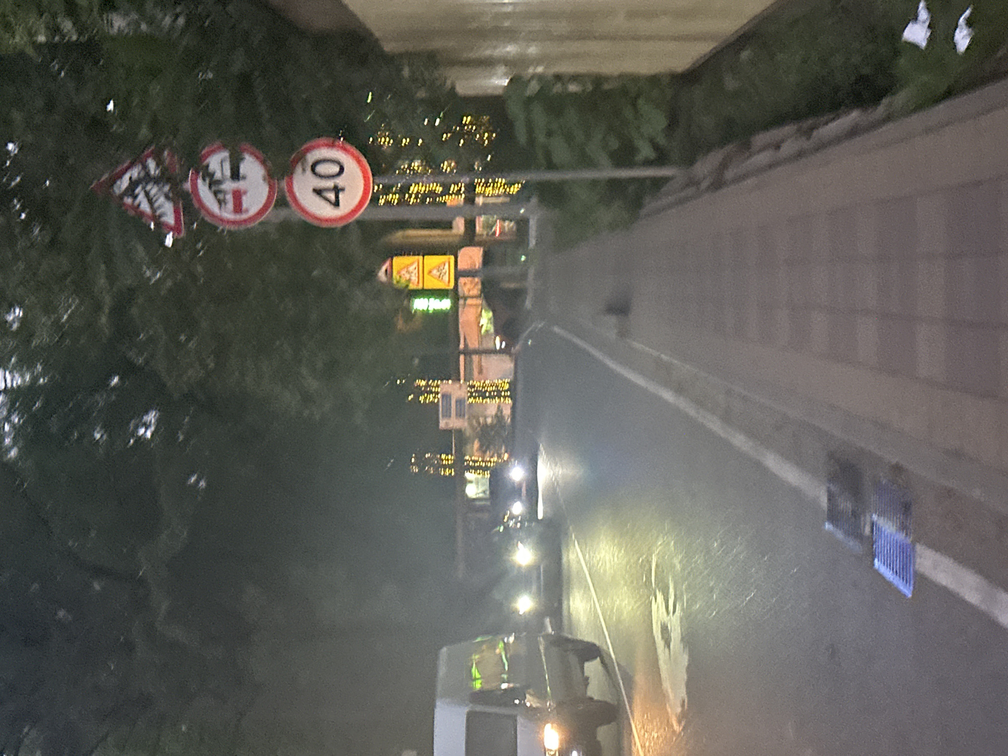 The image depicts a street scene at dusk or night in Sochi, Russia. A white van is visible on the left side of the road, with its headlights on. Several other vehicles are present further down the road, also with their headlights illuminated. The road is wet, suggesting recent rain. A sidewalk runs alongside the road, with a metal pole holding several traffic signs. The signs include a speed limit sign indicating "40", a sign prohibiting trucks, and a triangular warning sign. Trees with lights strung around them are visible in the background, contributing to a festive or decorative atmosphere. A building with lights is also visible in the distance. The overall impression is that of a city street at night, possibly in a commercial or entertainment district, given the decorative lighting.