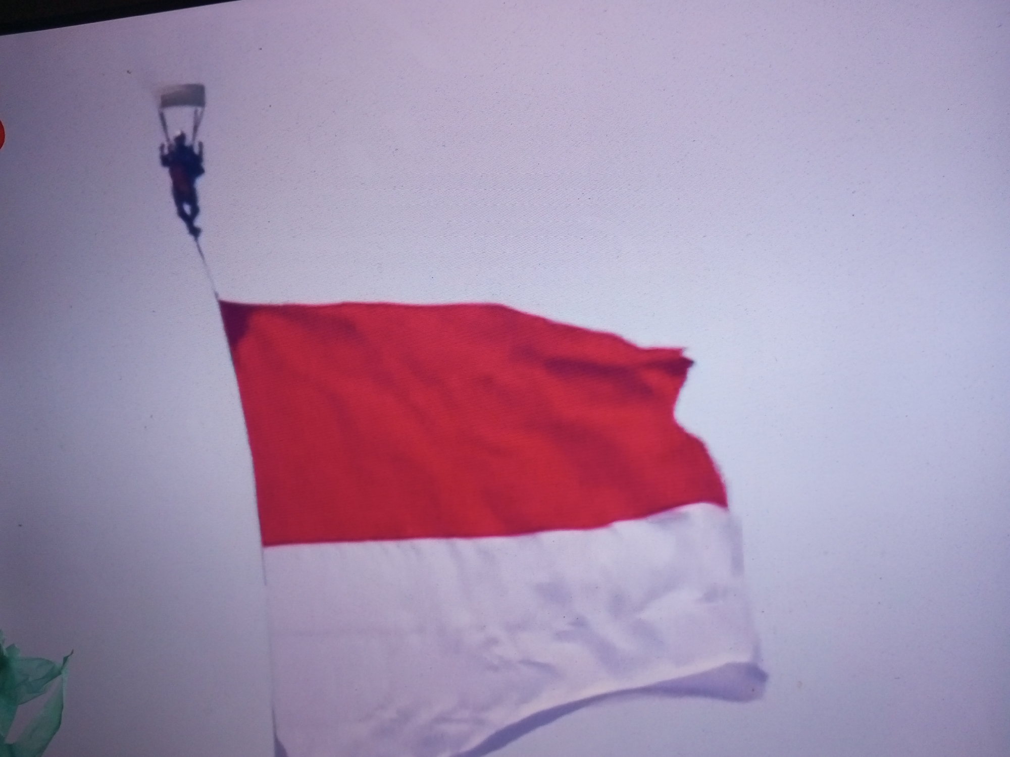 A skydiver descends with a parachute and appears to be holding a flag, possibly the Indonesian flag, which is red and white. The skydiver is positioned in the upper left quadrant of the image, above a large red and white flag that is billowing in the wind. The background is a light, overcast sky. The image quality is poor, with a noticeable grain and blur. No specific location in Banyudono, Indonesia, is identifiable from the image itself, as it is a generic depiction of a skydiver and a flag against a sky.
