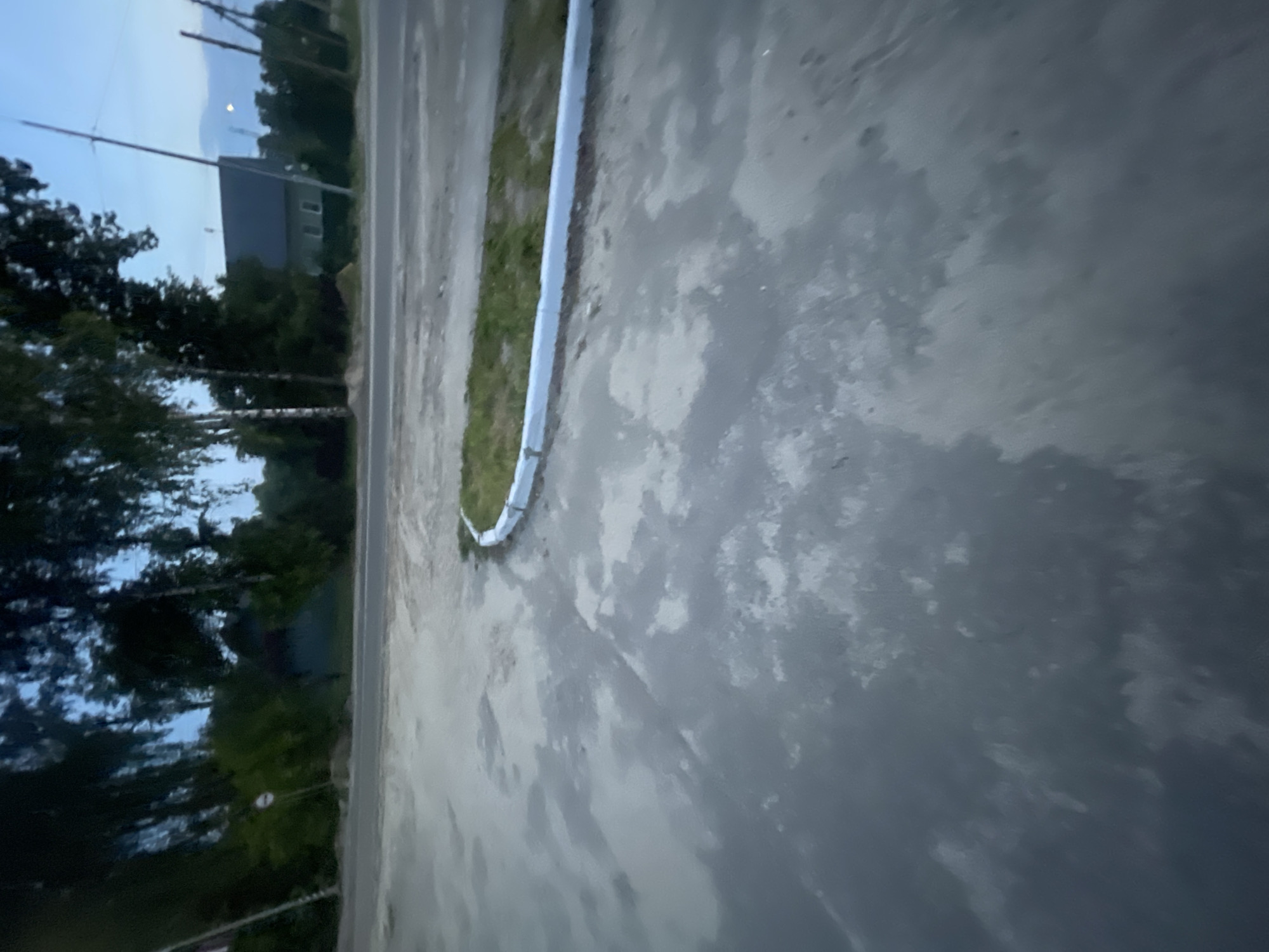 The image depicts a street scene in Krasnye Baki, Russia, likely during twilight. In the foreground, a wet, grey asphalt surface dominates the frame, reflecting the dim light. A curved strip of grass with a white border separates the asphalt from a road in the midground. Beyond the road, a residential building with a dark roof and several windows is visible, nestled amongst lush green trees. The sky above is a pale blue, hinting at the end of the day. Several utility poles are scattered across the landscape, some with visible wires. The overall impression is one of a quiet, rural setting.