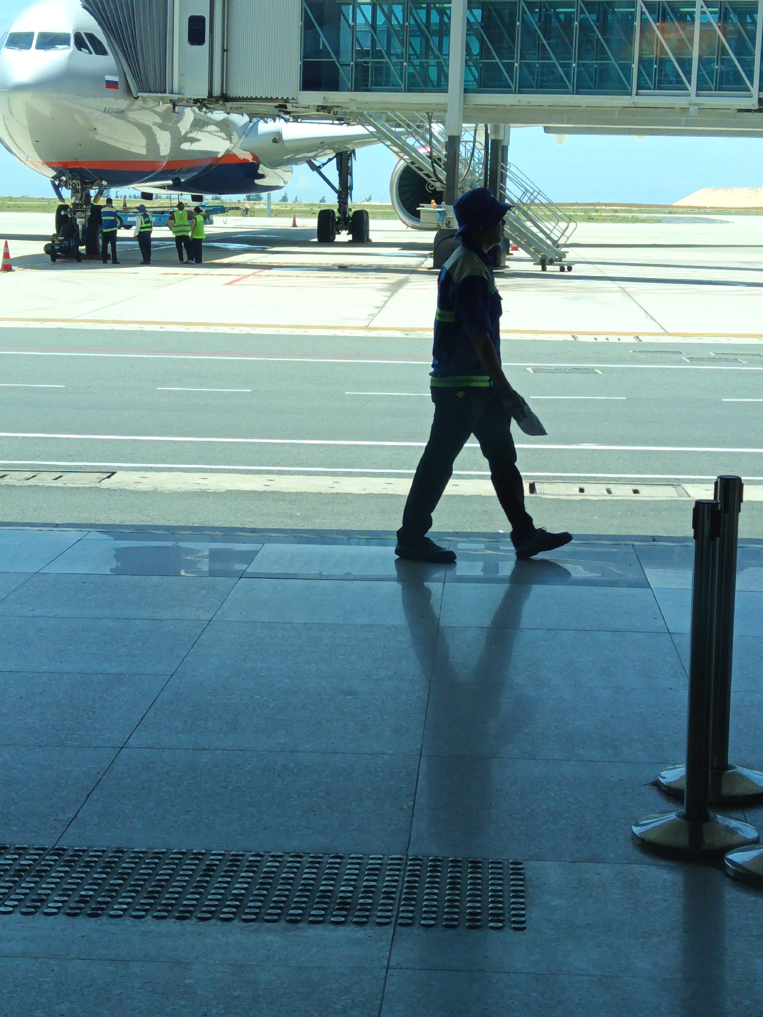 An airport scene in Bắc Cam Ranh, Vietnam, shows a commercial airplane connected to a jet bridge. In the foreground, a male airport worker wearing a dark blue uniform with green accents and a blue cap walks across a tiled floor. He is carrying a white document in his right hand. The worker's shadow is cast on the floor. Behind him, an airplane is visible, with its jet bridge extended to the aircraft. Several other workers in high-visibility vests are standing near the airplane's wheels. The environment appears to be an airport tarmac under a bright, clear sky.