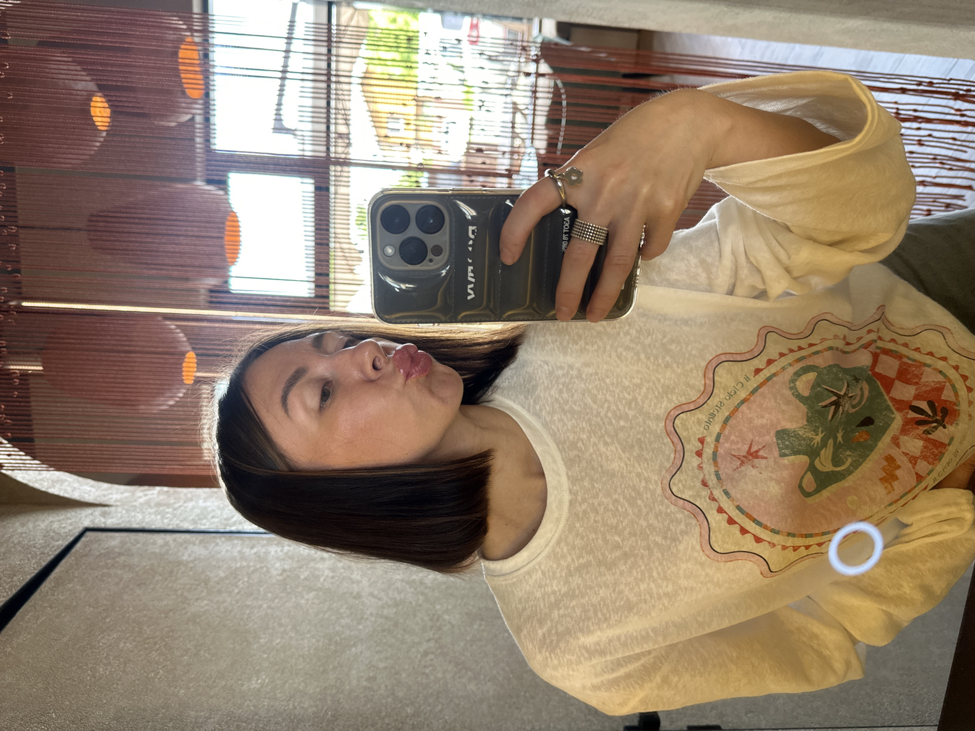 A young adult female is captured in a mirror selfie indoors, appearing to be within a commercial establishment such as a restaurant or cafe. She has medium-length, dark brown hair styled in a bob cut and is pouting her lips in a "duck face" expression, looking slightly downward and to her right. Her skin appears fair, and her lips are glossy. She is wearing a loose-fitting, off-white, long-sleeved t-shirt made of a textured (slub knit) fabric. The front of her shirt features a large graphic design within a scalloped, colorful frame. The central motif includes a stylized green teapot or pitcher, possibly with a spout resembling a bird's head, surrounded by what appears to be leaves, stars, and a checkered pattern in shades of red, white, and brown. Partial text is visible on the graphic, including "il cielo stellato" (Italian for "the starry sky") and "DI CIOCO," along with other illegible script. A small, bright circular reflection is visible on the lower left portion of her shirt. A sliver of light green or khaki fabric, possibly trousers or a skirt, is visible at the bottom edge of the frame. The woman holds a dark, possibly black or dark grey, padded or quilted smartphone case with her left hand, which holds the phone angled towards her face. The phone itself appears to be an iPhone model, identifiable by its triple rear camera array. Reversed text on the case reads "WANG" (suggesting Alexander Wang) and other partially visible, reversed text such as "DO SANTI" or "PRO 81 TOEA." Her left hand displays two rings on her thumb: a wide, multi-banded silver-toned ring, and another on her ring finger, which includes a simpler band ring and a charm ring with a small, silver-toned flower. The background reveals a stylish interior with a textured, light beige or grey wall on the left, featuring a dark vertical trim or doorframe. Further back, two arched architectural elements are visible. These arches are partially obscured by a screen or partition made of numerous reddish-brown vertical strings, beads, or thin fabric strips, creating a semi-transparent barrier. Within this screen, several large, spherical or bell-shaped light fixtures, also reddish-brown and translucent, are suspended, emitting a warm, orange glow. Beyond the screen and arches, large windows show an outdoor scene under bright daylight. Green foliage, trees, and parts of residential or commercial buildings (one with a light-colored roof) are discernible