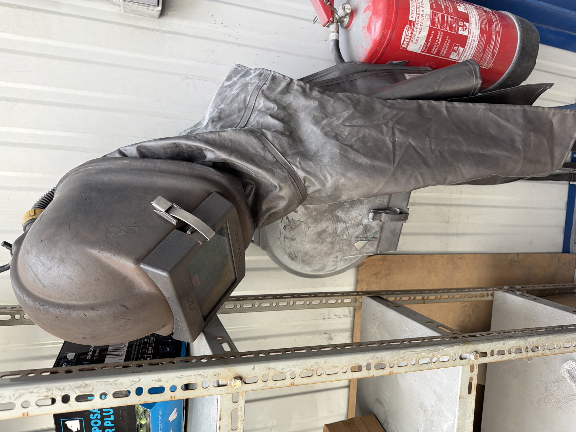 In Mol, Belgium, a welding helmet and protective suit hang on a metal rack next to a fire extinguisher. The scene appears to be an indoor storage area, possibly a workshop or industrial space, with a corrugated metal wall and industrial shelving units visible. The welding helmet is dark gray and appears to be a professional-grade model, with a visible visor and straps. The protective suit is also dark gray and made of a heavy-duty material, designed to shield the wearer from sparks and heat. The fire extinguisher is red and appears to be a standard industrial model. The overall impression is one of organized safety equipment, ready for use.