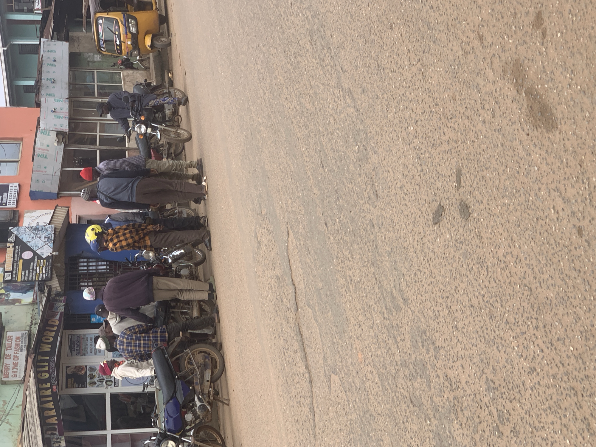 The image depicts a street scene in Shagamu, Nigeria. In the foreground, a section of an asphalt road is visible, showing signs of wear and tear with some patches and cracks. On the right side of the road, a yellow auto-rickshaw, commonly known as a "Keke Napep," is parked.
Behind the road, a row of buildings lines the street. The most prominent building in the mid-ground features signage that reads "ARAIRE GLIT WORLD" with a phone number. To the left of this shop, a blue motorcycle is parked on the side of the road. Further to the right, several men are gathered. Some are standing next to motorcycles, with one individual seated on a motorcycle. The men are wearing casual clothing, including shirts, trousers, and some are wearing caps or helmets. One man is wearing a yellow helmet, and another has a red beanie. Behind this group, more motorcycles are visible.
The buildings show various architectural styles and states of repair. Some appear to be shops, with windows and entrances facing the street. One building has a bright orange facade. Signs in English are visible on the buildings, indicating businesses such as "MERRY DE TAILOR" and "GHOME OF FASHION." The overall impression is of a bustling urban environment, likely a commercial area within Shagamu.