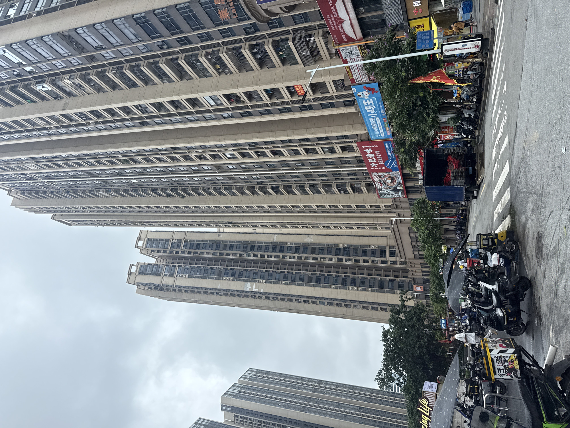 In Haizhu District, China, this image captures a street scene with several tall residential buildings dominating the background. The buildings are modern, with numerous balconies and windows. In the foreground, a street is visible, wet from recent rain, with a pedestrian crossing. Parked on the side of the street are numerous motorcycles and scooters, some covered with tarps. A blue truck is also parked on the street, its rear doors open, revealing bags of what appear to be red tiles or bricks. A few people are visible in the scene, some standing near the motorcycles, one walking on the sidewalk, and another near the truck. Street banners and advertisements are present on the buildings and poles. The sky is overcast, suggesting a cloudy day.