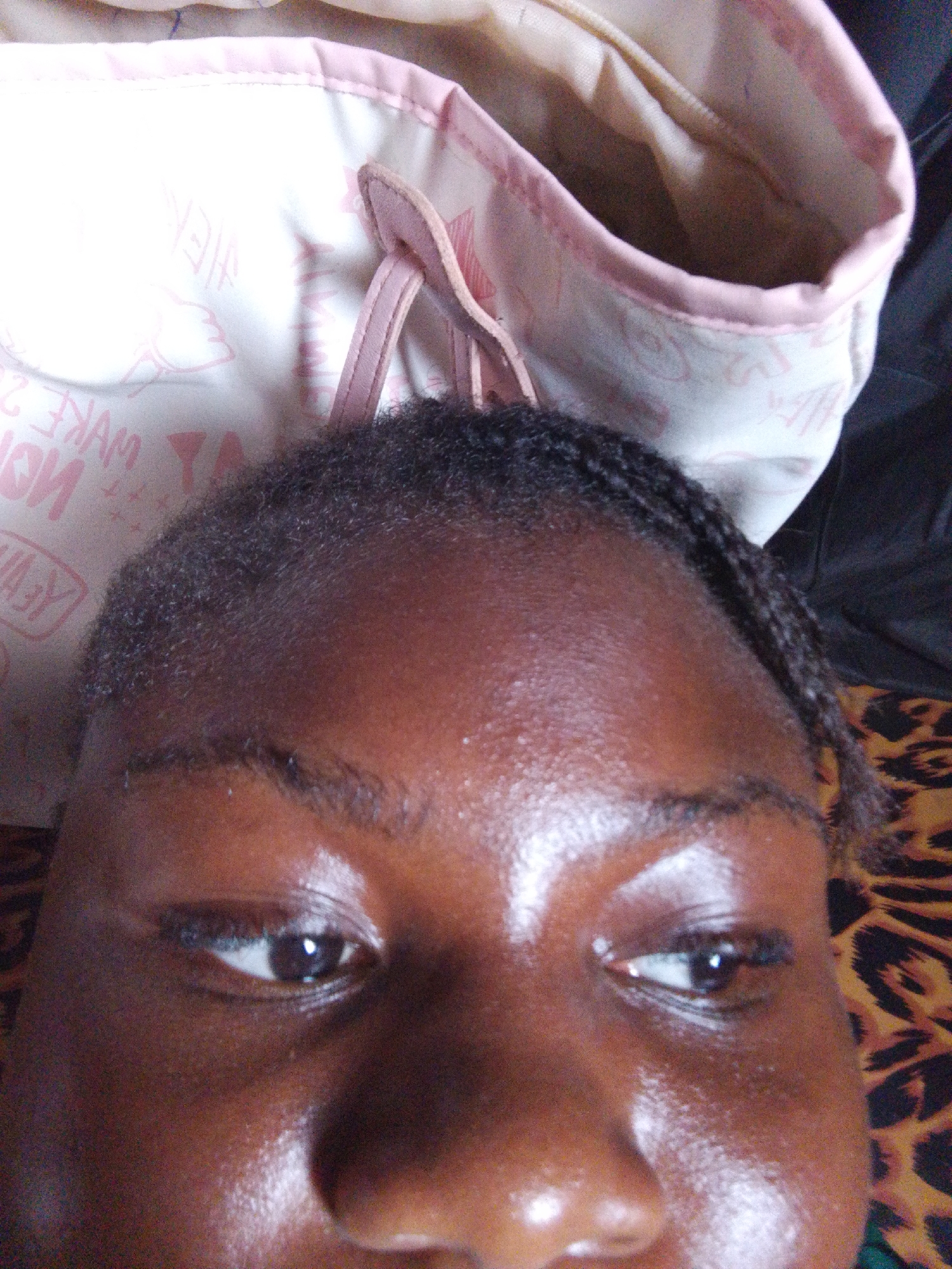 The image presents a close-up, low-angle shot of a person's upper face, encompassing their forehead, eyes, and nose. The individual has a dark skin tone, with a noticeable sheen on their skin, particularly across the forehead, nose, and cheeks, suggesting either an oily complexion, perspiration, or bright direct lighting. Their dark eyes are open and appear to be looking slightly upwards. Dark, well-defined eyebrows frame the eyes. The hairline shows natural, dark, tightly curled hair, with a section of neat braids visible along the right side of the head.
In the upper background, a light pink and white fabric bag or pouch is partially visible. It features thin pink straps and is adorned with repeating pink text and graphic designs. The text is mirrored, with discernible words like "MAKE" and possibly "UP" or "24" towards the lower left, and "HEY" or "HELLO" higher up, all rendered in pink against the white fabric. To the right and below the person's head, a segment of brown and black leopard print fabric adds a contrasting pattern to the background.
The overall lighting appears bright, creating significant highlights on the skin. The image conveys a close-up, personal perspective, likely a selfie or an intimate portrait. The location context for this image is Nsukka, Nigeria, although no specific visual cues within the frame directly indicate this geographical setting.