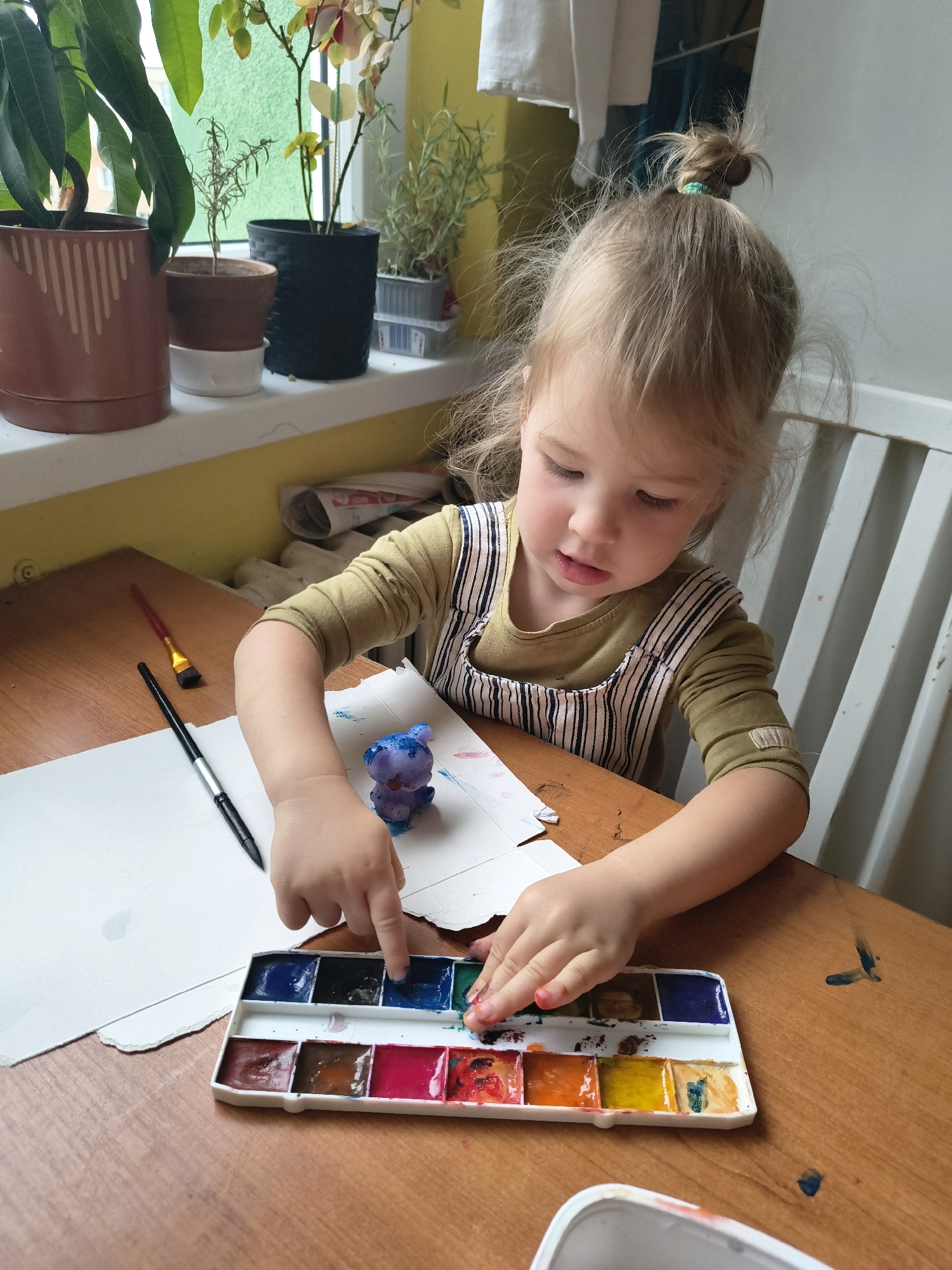 A young child, with blonde hair pulled into a messy bun, is focused on painting at a wooden table. They are wearing a long-sleeved olive green shirt and striped overalls. In front of them is a watercolor paint palette with various colors, some of which have been used. The child's fingers are dipped into a blue paint compartment. To the left of the palette, there is a piece of white paper with some paint smudges and a small purple figurine that appears to be painted. Two paintbrushes are also on the table, one black and one with a red handle and gold bristles. The background shows a windowsill with several potted plants. The setting appears to be an indoor domestic environment, possibly a home in Olomouc, Czechia, indicated by the context provided.