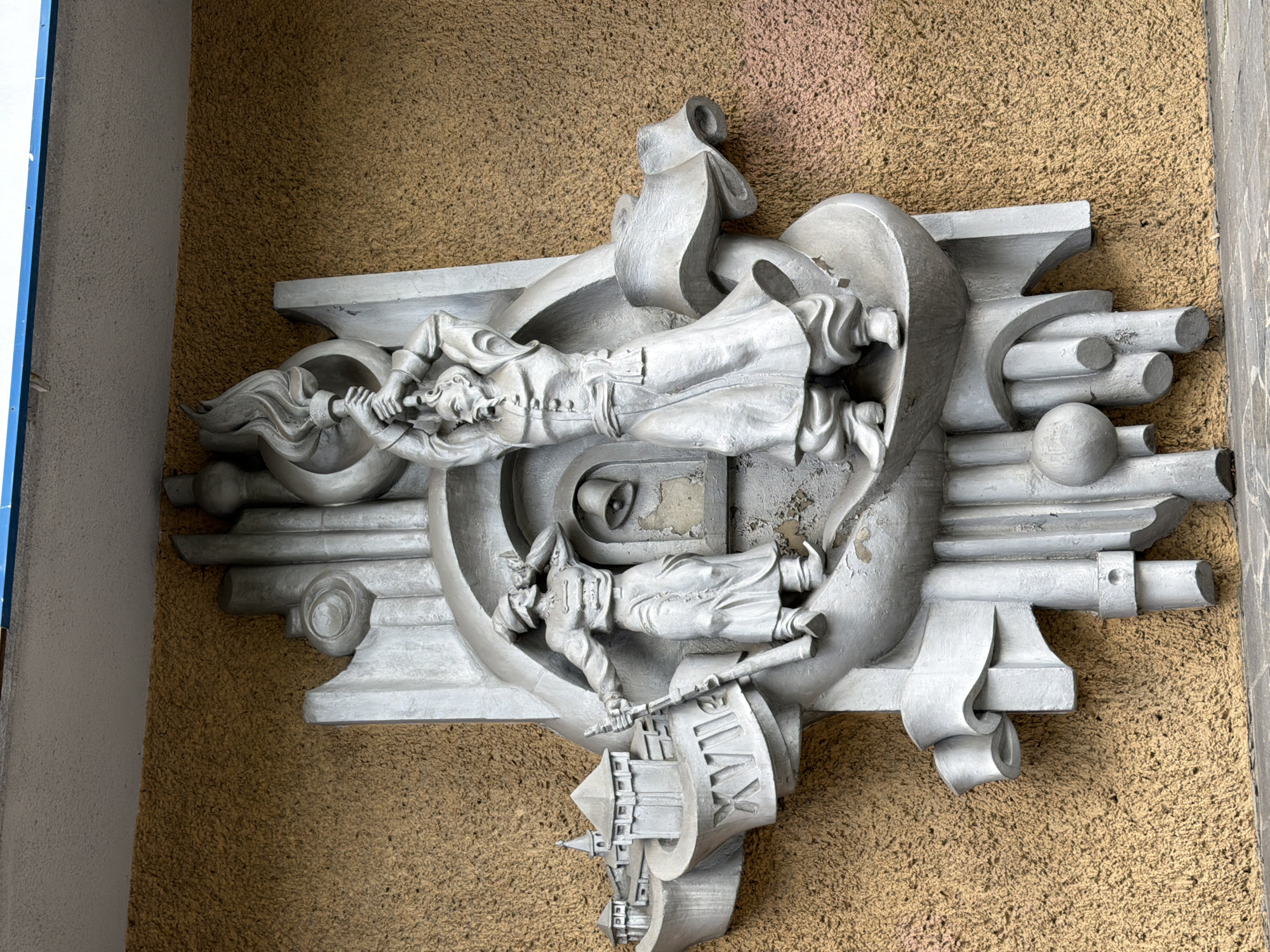 This is a detailed, low-angle, full-shot photograph of a large, gray, relief sculpture mounted on a textured, mustard-yellow building facade. The sculpture, which appears to be made of concrete or a similar material, is predominantly silver in color. It depicts two figures, likely historical Cossacks based on their attire, in a dynamic composition.
The left figure, positioned on the left side of the sculpture, wears a traditional Cossack hat and military-style tunic, with a sash around his waist. He holds a long rifle vertically in his right hand, with his left arm raised to his head, perhaps signaling or in a defensive stance. To his left, a stylized representation of a fortress or town with spires and crenellations is visible, alongside a banner bearing the Roman numeral "XVII".
The right figure, larger and more prominent, stands in the center, looking upwards. He wears a similar hat and tunic, with a sash and wide trousers. He is depicted raising a flaming torch high above his head with both hands, symbolizing enlightenment or revolution. His expression is one of intensity and determination.
Beneath the figures, the sculpture features abstract elements, including curves, cylindrical shapes, and circular forms. A bell is visible in the background behind the left figure. The overall composition suggests a historical narrative related to the 17th century, possibly referencing a significant event or period in Ukrainian history. The building facade is rough and textured, with some areas showing peeling paint, particularly around the bell in the sculpture. The upper edge of the image shows a blue overhang and a white soffit. The ground in the foreground consists of stone paving slabs.
This sculpture is located on the facade of the Zhytomyr Regional State Administration building in Zhytomyr, Ukraine.