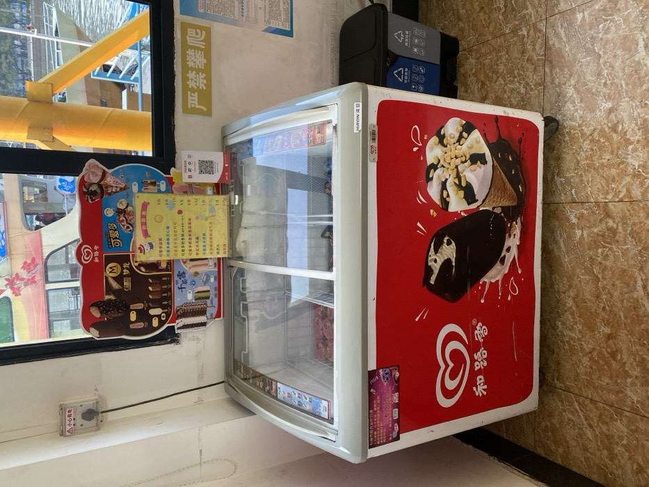 The media file, rotated clockwise by 90 degrees, captures an indoor scene, likely a small shop or kiosk in Guangzhou City, China, situated next to a body of water.
The main subject is a white ice cream display freezer with a glass top, featuring a prominent red side panel adorned with large illustrations of ice cream cones and bars, alongside the "Wall's" (和路雪) brand logo and a heart icon. The word "HIRONB" is also visible on the freezer's top edge. Attached to the freezer, on the left, is a colorful menu board displaying various ice cream options, including "可爱多" (Cornetto) and "梦龙" (Magnum), with prices listed in Chinese Yuan (e.g., ¥4, ¥6, ¥8, ¥12, ¥16, ¥18). A QR code is present on the menu board, likely for payment or information. The glass display section of the freezer appears mostly empty.
Behind the freezer, a black recycling bin with separate compartments and Chinese text for waste categories is visible. To the left of the freezer, a window offers a view of a body of water and a yellow structure, possibly part of a boat or a railing by the waterfront. On the wall above the freezer, a yellow sign with red Chinese characters reads "严禁攀爬" (Strictly forbidden to climb). Below the window, a small white electrical box has a warning label with "▲小心有电" (Caution: Electricity). The floor is tiled in a light brown, marble-like pattern.
The bright lighting suggests it is daytime. The presence of the ice cream freezer, menu, and waterfront view indicates a retail point, possibly catering to visitors or locals in a recreational area.