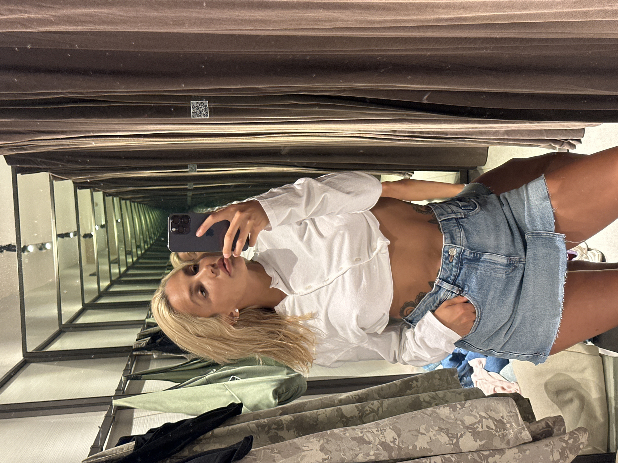 In Kuta, Indonesia, a young woman with blonde hair takes a mirror selfie in what appears to be a fitting room or boutique. She is wearing a white, cropped long-sleeved shirt and a light blue denim skirt. Her right hand is in her skirt pocket, and a tattoo is visible on her hip. The background shows clothing racks with various garments, as well as rows of floor-to-ceiling mirrors that create a hallway effect. To the right, a brown curtain is visible, with a QR code attached to it. The lighting suggests an indoor setting, possibly a retail store.