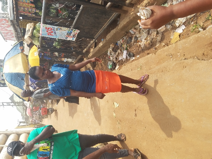 The image depicts an active street market scene in Nnobi, Nigeria, under daylight conditions.
In the mid-foreground, a woman, approximately 30-40 years old, is walking towards the viewer's left. She wears a blue, short-sleeved top with a white undergarment visible at the neckline, an orange knee-length skirt, and purple-striped flip-flops. Her hair is dark and short. She looks downwards and to her left.
To the woman's immediate right, an open drainage ditch or gutter runs parallel to the walking path, filled with accumulated refuse including plastic bags, wrappers, and various debris. The walking path itself is a light brown, packed dirt or concrete surface, showing a distinct shadow of a person in the foreground.
To the left of the primary subject, a man, approximately 30-50 years old, walks towards the right. He wears a green t-shirt with a colorful graphic print, dark trousers, and a black baseball cap with a white "NY" logo. He holds a white cloth or tissue to his face with his right hand. A partially visible leg and foot, wearing dark sandals, are present in the far left foreground.
Behind the woman and separating the pathway from the market stalls is a black metal fence. A white rectangular poster advertising "POS ISHALALALE" is attached to this fence. Beyond the fence, several market stalls are visible. One prominent stall is shaded by a blue and white striped umbrella, while another has a yellow umbrella. Piles of green leafy vegetables are displayed on a surface. A sign above one stall reads "POSPOS".
In the background, further down the street, more pedestrians, including a bald man in a grey t-shirt, and a motor scooter are visible amidst additional market structures. Utility poles and overhead wires are present against a bright, hazy sky. The overall environment indicates a bustling commercial area with visible waste accumulation along the drainage.