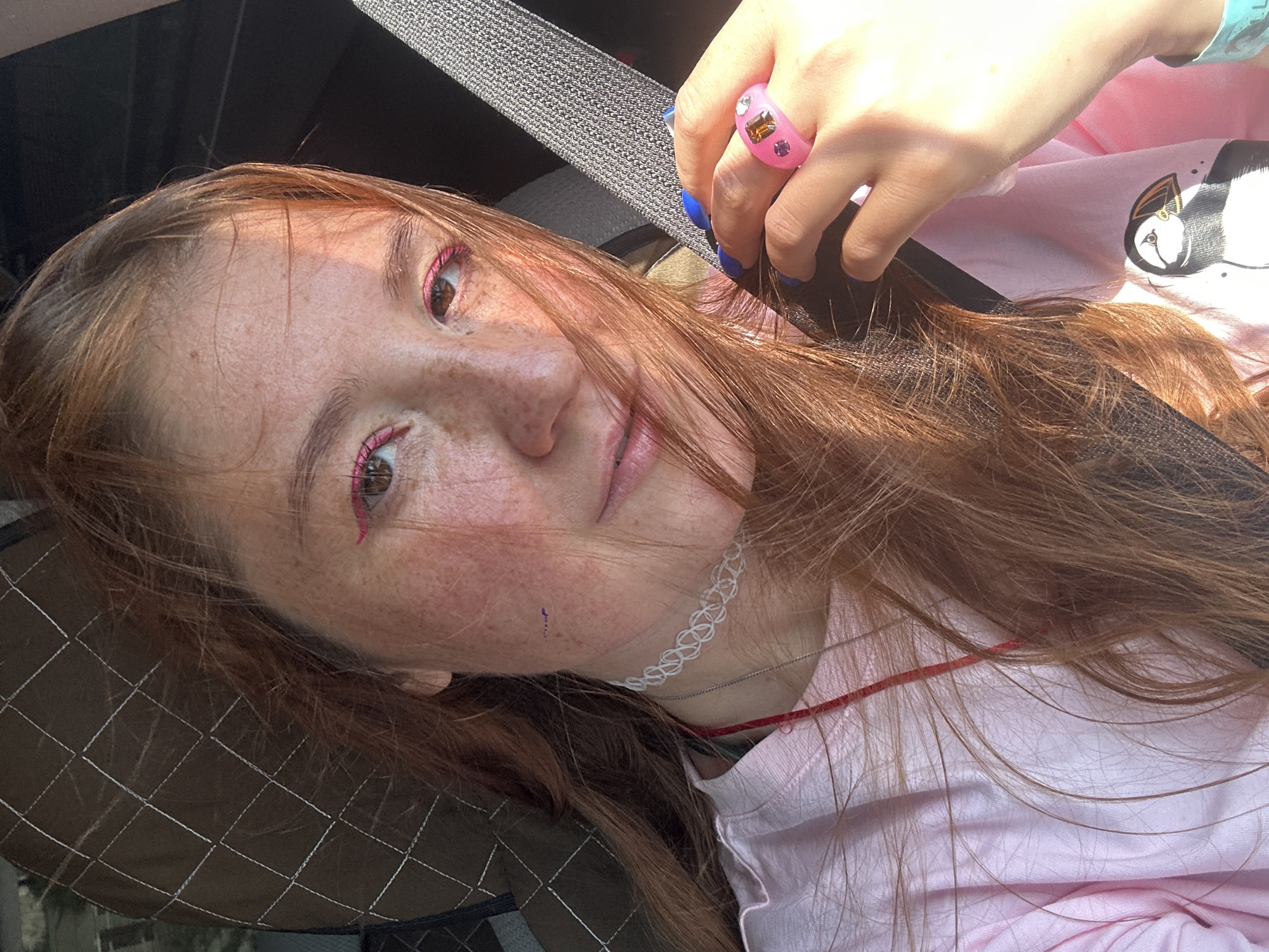 In this selfie, a young woman with red hair and freckles is seen from the chest up. She is wearing a pink t-shirt with a puffin graphic on it and a white choker necklace. Her makeup includes bright pink eyeliner. On her right hand, she wears a pink ring with gemstones and her nails are painted blue. The background shows a car interior, with a seatbelt and part of a headrest visible. The lighting suggests it is daytime, possibly with sunlight filtering through. The location is not specified as Крутобереговый, Russia, but the image is a close-up selfie taken within a vehicle.