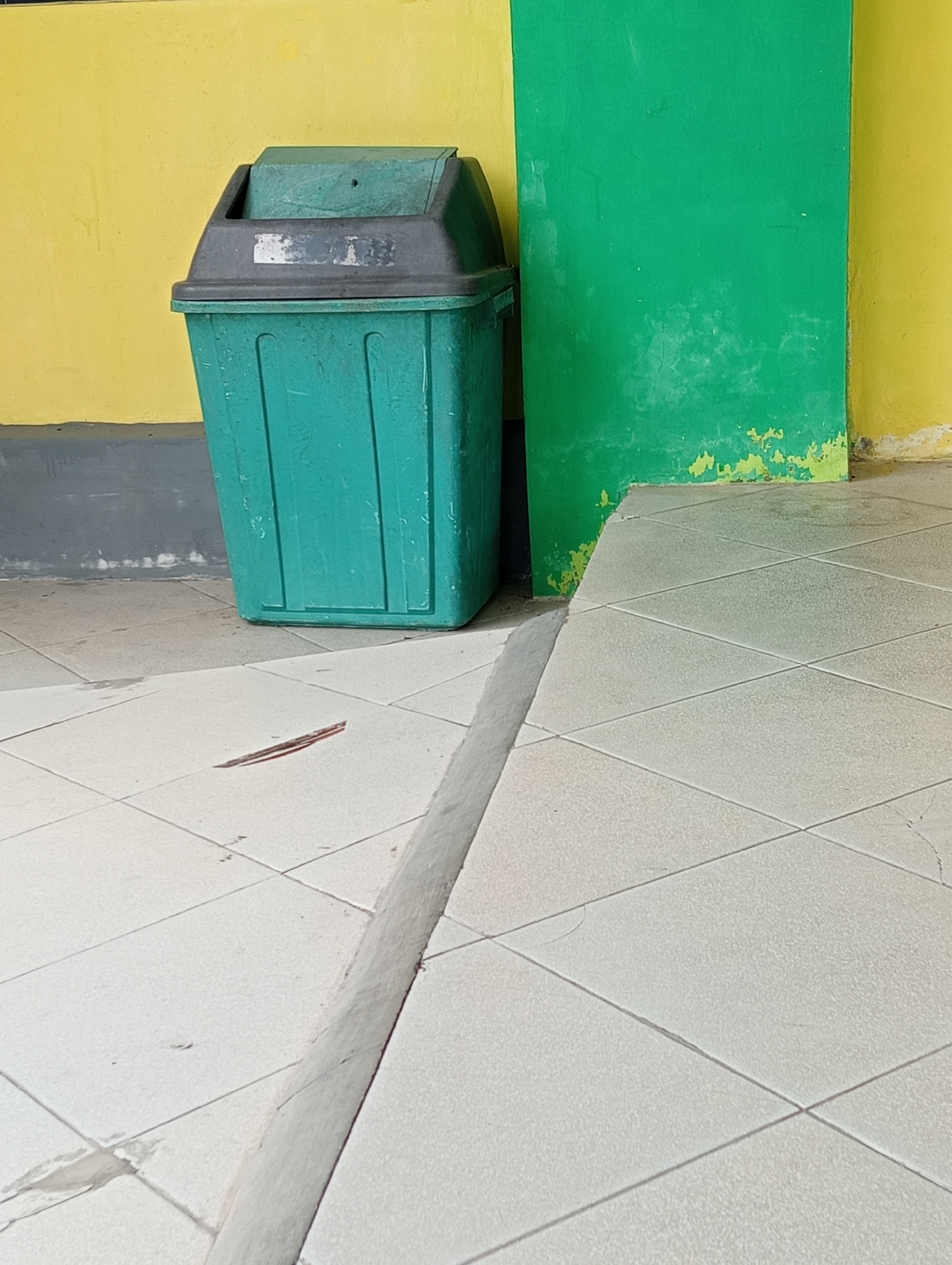 A green trash can with a black lid is positioned against a yellow wall. To the right of the trash can is a green wall. The ground is tiled with light-colored squares. A raised concrete strip runs across the foreground. A small red and brown object is visible on the tiles in the lower left quadrant. The scene appears to be an outdoor public space, possibly in Sidoharjo, Indonesia, characterized by utilitarian construction and weathered surfaces. No people or animals are present.