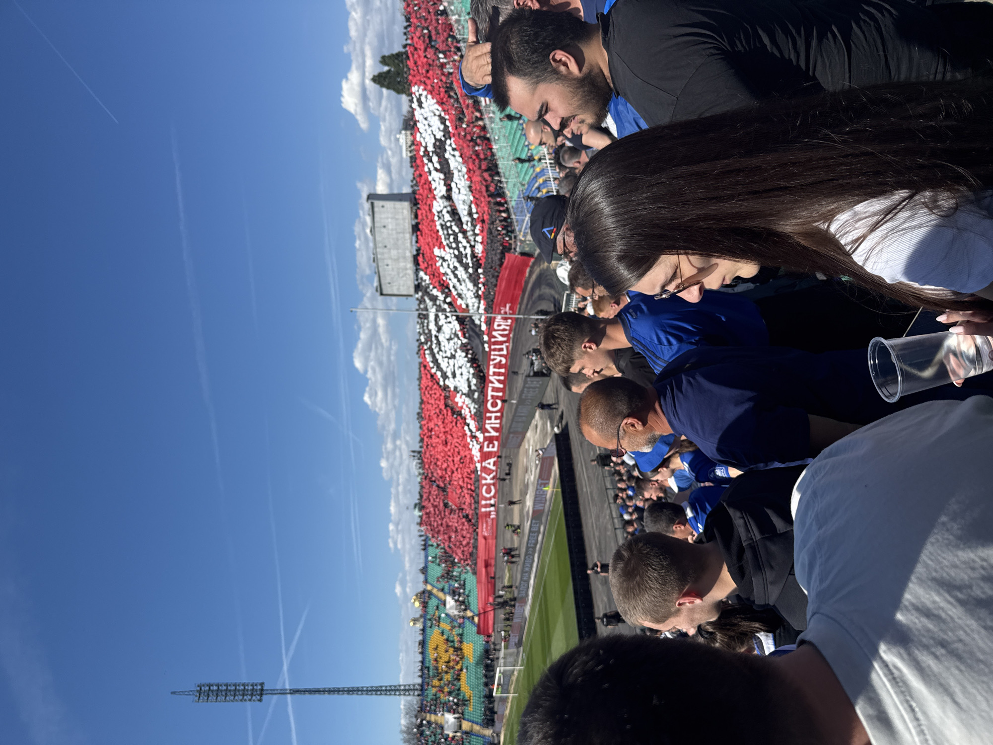 A tilted eye-level outdoor shot shows a crowd of people attending a sporting event in Unknown, Unknown. The event is possibly a soccer match, given the green playing field visible in the middle of the frame, flanked by spectators in the stands. A large banner with red, white, and black coloring stretches across the upper section of the stands, with legible lettering on the banner that reads "ACKA E VHCTHT YUHH." The people in the foreground appear to be spectators as well, wearing casual clothing in various shades and colors. A plastic cup is seen in the right side of the frame. A clear blue sky with a few scattered clouds is visible above the stadium. Tall light posts are located on the left of the frame.