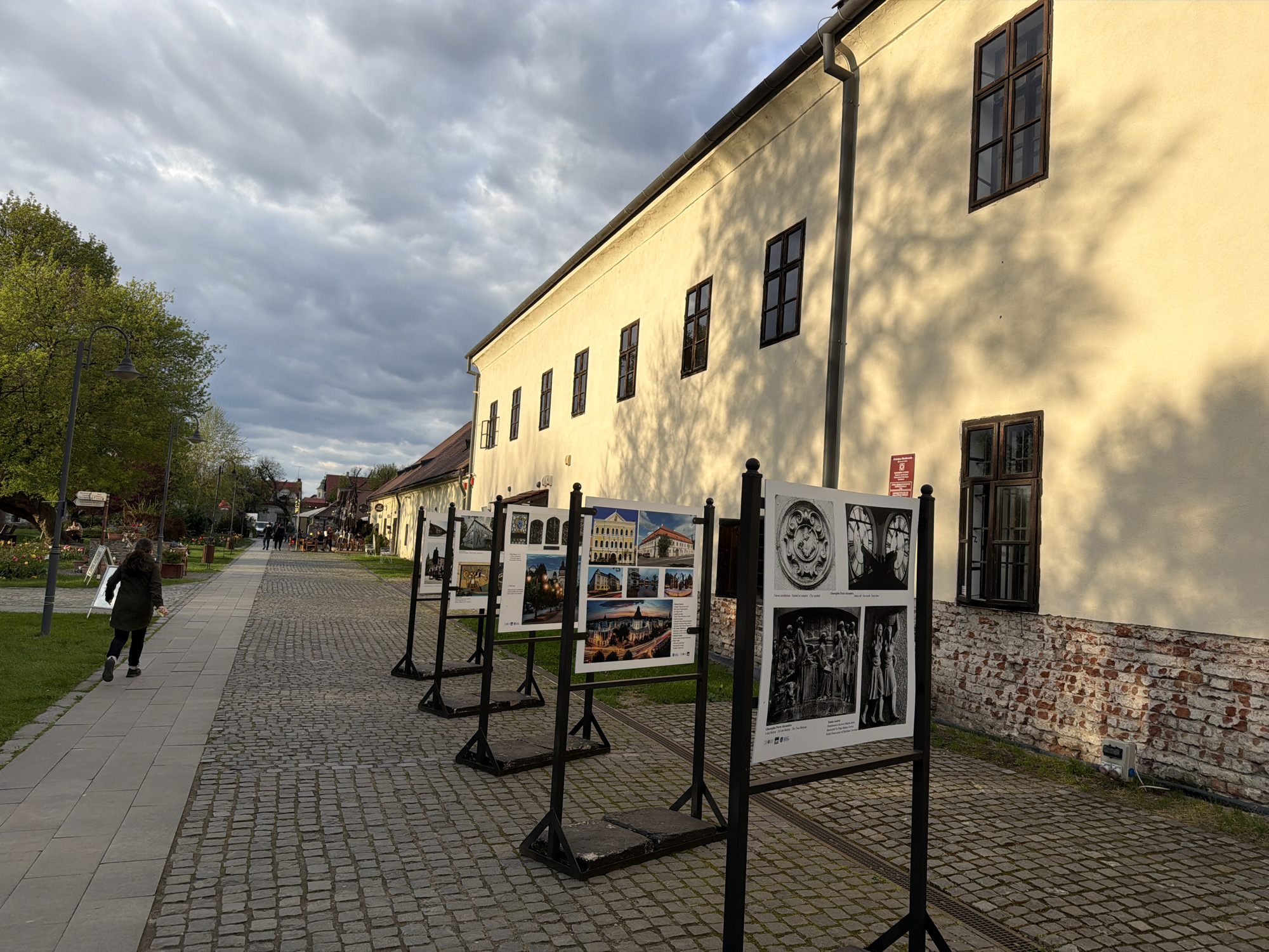 An outdoor photography exhibition is displayed along a cobbled street in Târgu Mureș, Romania, likely during the late afternoon or early evening due to the long shadows cast. The sky is overcast with a textured cloud cover.
The scene features a long, light yellow building with multiple dark-framed windows, against which the shadows of nearby trees are distinctly projected. A gray drainpipe runs down the side of the building, and a section of its lower wall reveals red brick. A small red rectangular sign with white text, possibly an official emblem, is affixed to the building's wall.
In the foreground, a paved sidewalk runs parallel to the cobbled street. On the sidewalk, a person wearing a dark jacket and trousers is walking away from the camera. Along the cobbled street, a series of metal stands hold large white panels, each displaying multiple photographs. These photos appear to depict architectural details, cityscapes, and historical scenes, possibly of Târgu Mureș. The panels have small text captions and what appears to be copyright information or credits, with words like "Fotografia" (Photography) and "Descrierea" (Description) discernible on some. One panel shows round architectural motifs, a clock face, and sculpted figures.
Further down the street, more people are visible in the distance, and the area suggests a pedestrian zone with trees, streetlights, and what look like outdoor seating areas for cafes or restaurants. The overall atmosphere is tranquil and inviting.