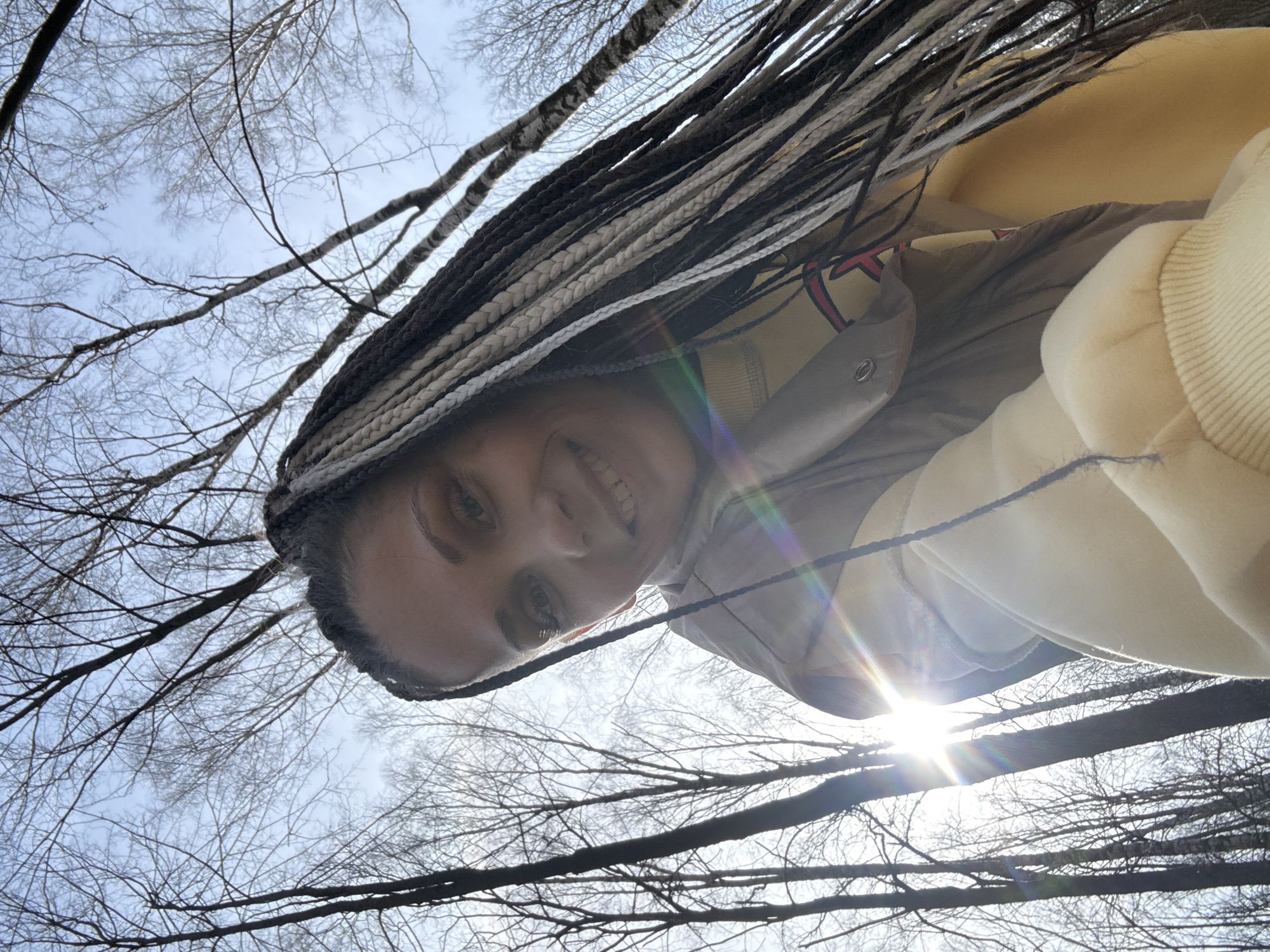 A low-angle selfie photograph depicts a person smiling at the camera, positioned against a backdrop of bare tree branches and a bright sky. The person has braided hair with a mix of dark and light strands, and wears clothing with tan and yellow tones. The sun is visible through the branches, creating a lens flare. The location is not provided in the image, therefore the city of Unknown, Unknown cannot be verified.