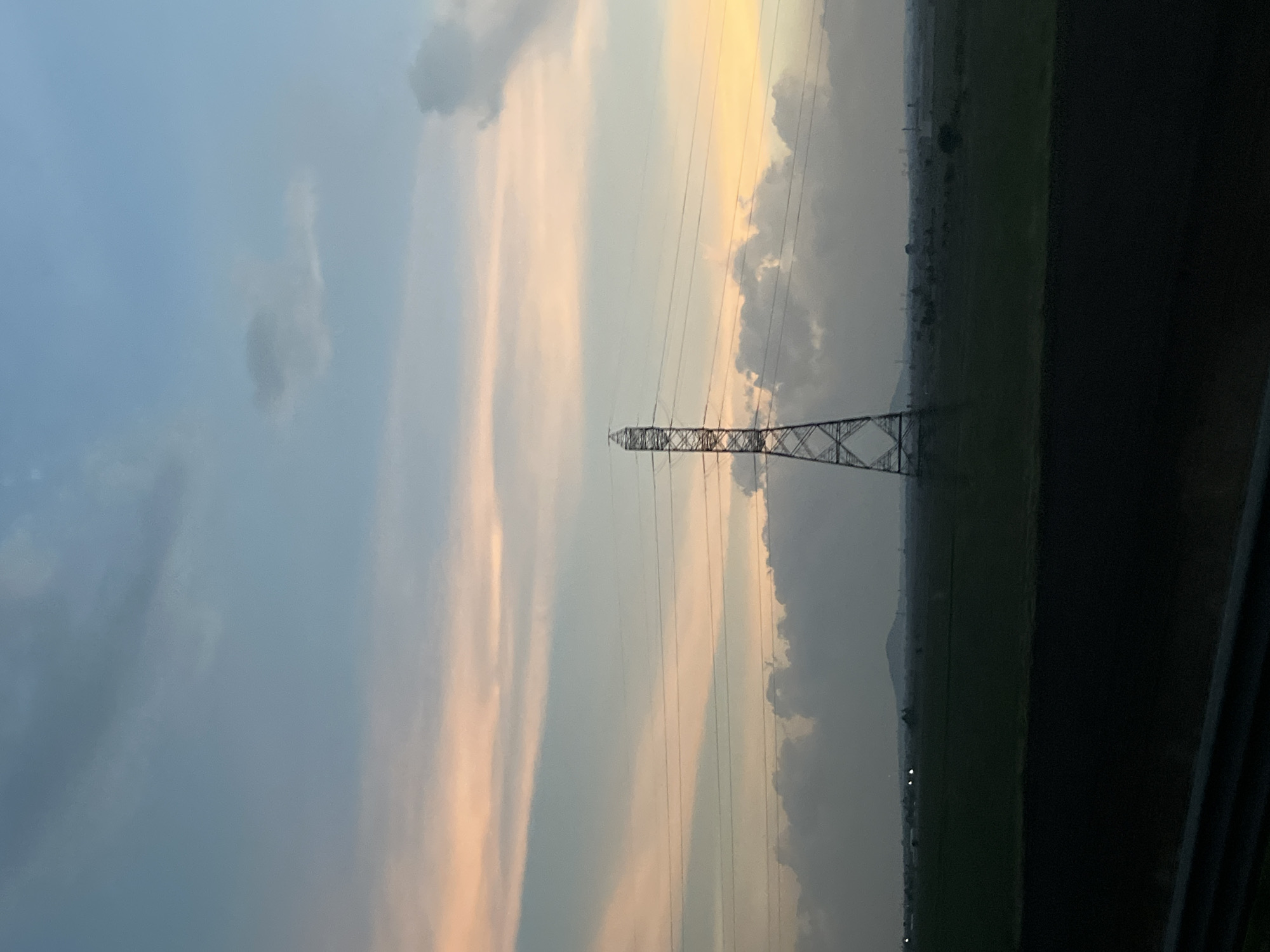The image depicts an electrical transmission tower standing in a field, likely agricultural, near Calpulalpan, Mexico. The sky behind the tower is filled with clouds, some reflecting the colors of the setting sun. There is a dark, solid barrier on the right side of the image.
