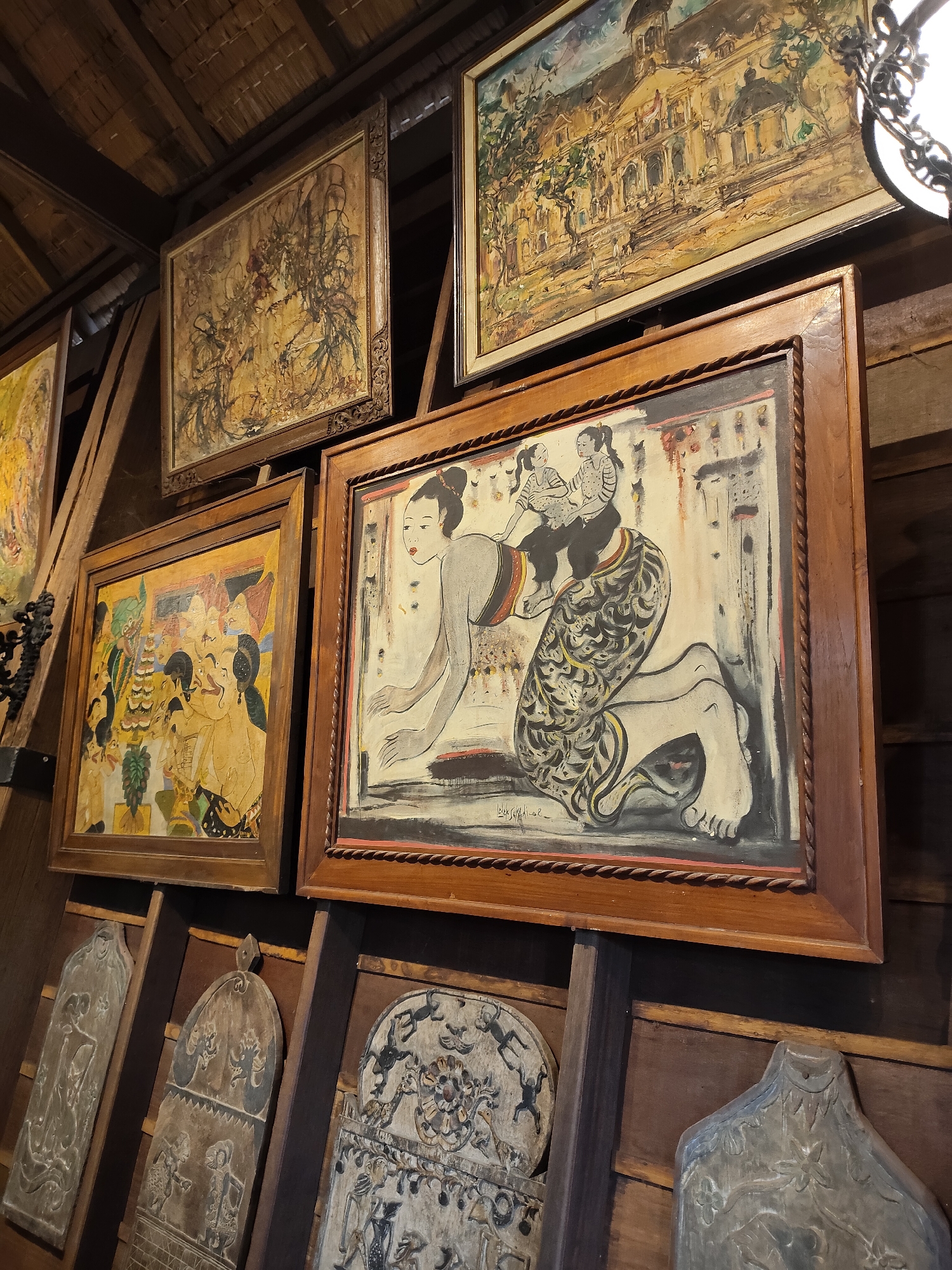 The image shows a collection of framed paintings and carved wooden plaques displayed on a wooden wall, possibly in an indoor setting. The paintings appear to be in a traditional style, with one central painting depicting a woman carrying a child on her back, and other paintings showing various figures and scenes. The carved wooden plaques are displayed below the paintings, featuring intricate patterns and figures. The overall impression is that of an art exhibition or a display of cultural artifacts. The context suggests that this scene might be located in the city of Medan, Indonesia, given the prevalence of traditional art forms in the region.