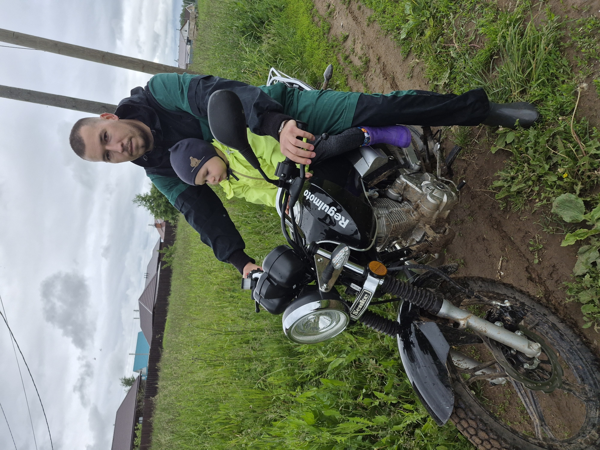 The image displays an adult male and a young child seated on a black "Regulmoto" motorcycle in a rural or semi-rural setting in Kharnavy, Russia. The motorcycle is positioned on a muddy dirt path, bordered by tall, unkempt green grass and scattered weeds.
The adult male, with short buzzed hair and light facial stubble, wears a black and dark green hooded jacket, matching dark green pants, and dark grey rubber boots. He is seated on the motorcycle, holding the handlebars with both hands, and looks directly at the viewer with a neutral expression. His left foot rests on the muddy ground.
A young child, likely a toddler, sits in front of the adult male on the motorcycle. The child is facing towards the left side of the frame, looking forward, and wears a neon yellow jacket, a dark grey beanie adorned with a light brown bear emblem, dark grey leggings, and bright purple patterned rubber boots.
The black motorcycle has the brand name "Regulmoto" visible on its fuel tank and "Senke" on the chrome bracket above the round headlight. The front wheel, fender, and lower engine components are splattered with mud. The motorcycle features knobby tires and a visible front disc brake.
The background shows an overcast sky with grey clouds, where three small, dark, distant airborne objects are faintly discernible. Two leaning wooden utility poles with overhead power lines stand behind the subjects. Further in the distance, a dark brown fence separates the field from several single-story houses with brown and blue roofs.