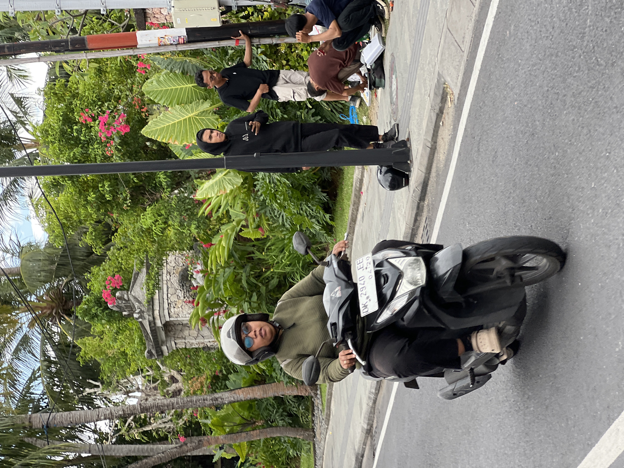 A woman wearing a green long-sleeved shirt, black pants, a helmet, and sunglasses rides a black scooter on a paved road in Denpasar, Indonesia. The road has a white line marking on the left side. Behind her, on the sidewalk, a group of people are gathered near a lamppost. One person in a black hoodie holds a phone, another in a black polo shirt and khaki shorts leans on the lamppost, and two individuals in the foreground are crouched down, appearing to be working on something on the ground. Lush green foliage and pink bougainvillea flowers are visible in the background, suggesting a tropical environment. A stone wall with decorative elements is also present.