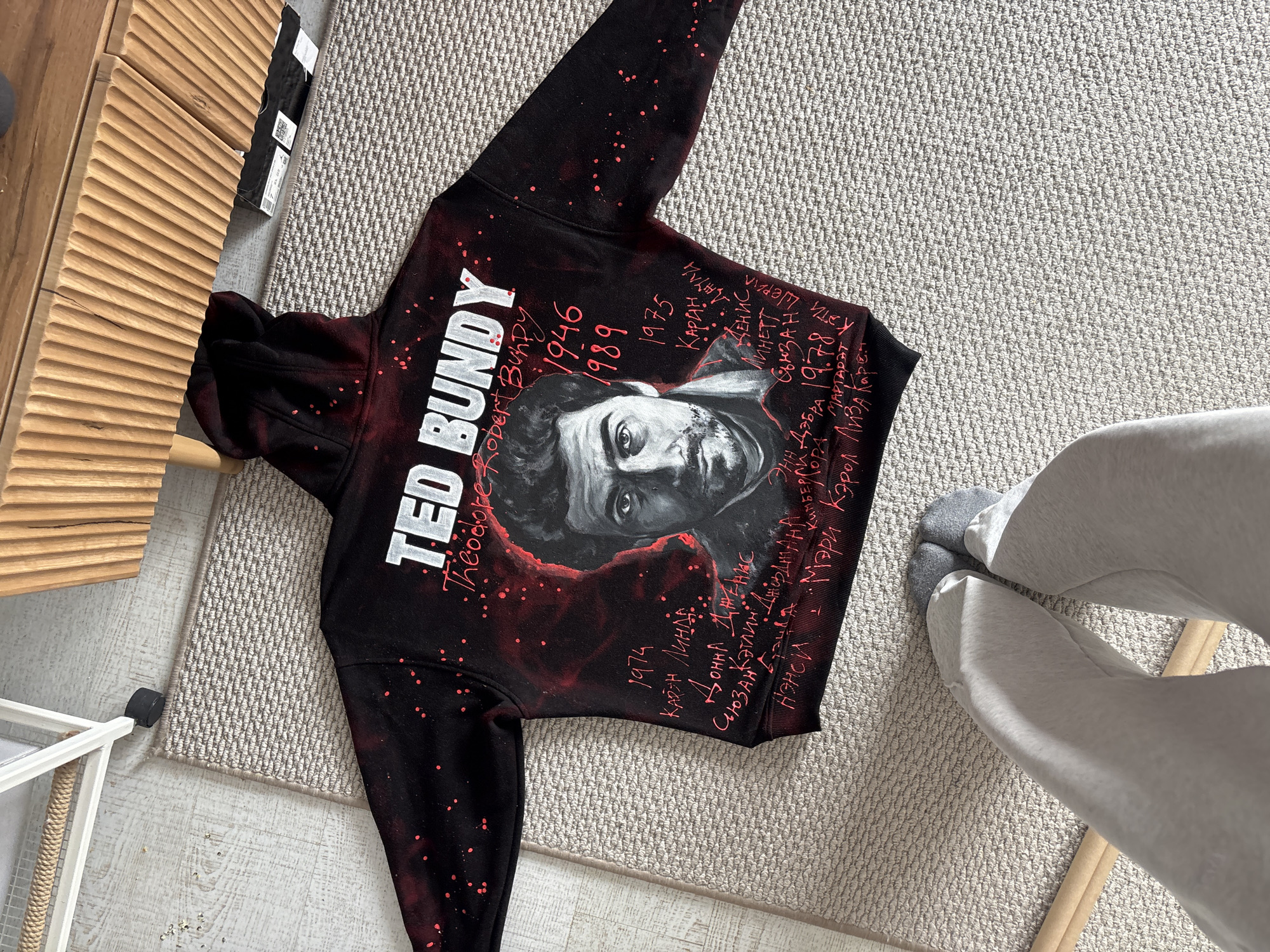 A black hoodie with a portrait of Ted Bundy is laid on a light grey rug. The hoodie has red splatter patterns and handwritten text in red. The text includes Bundy's name, birth year (1946), and execution year (1989), along with a list of names and the year 1974. Parts of a person's legs, clad in light grey sweatpants and grey socks, are visible in the bottom right corner of the image. The location is not in Krasnoyarsk, Russia. Other background elements include a wooden cabinet and a white wire rack.