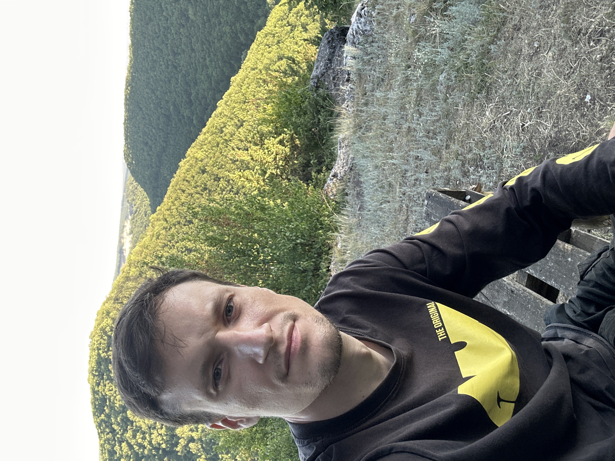 A man with short brown hair and stubble is sitting on a wooden bench in an outdoor setting. He is wearing a black long-sleeved shirt with a yellow graphic of a bat symbol and the text "THE ORIGINAL" on the left chest. The man is looking towards the camera. The background consists of rolling, tree-covered hills bathed in sunlight, with some dry grass and rocks in the foreground. The location appears to be a natural landscape, possibly on the outskirts of Pryvorottia Druhe, Ukraine.