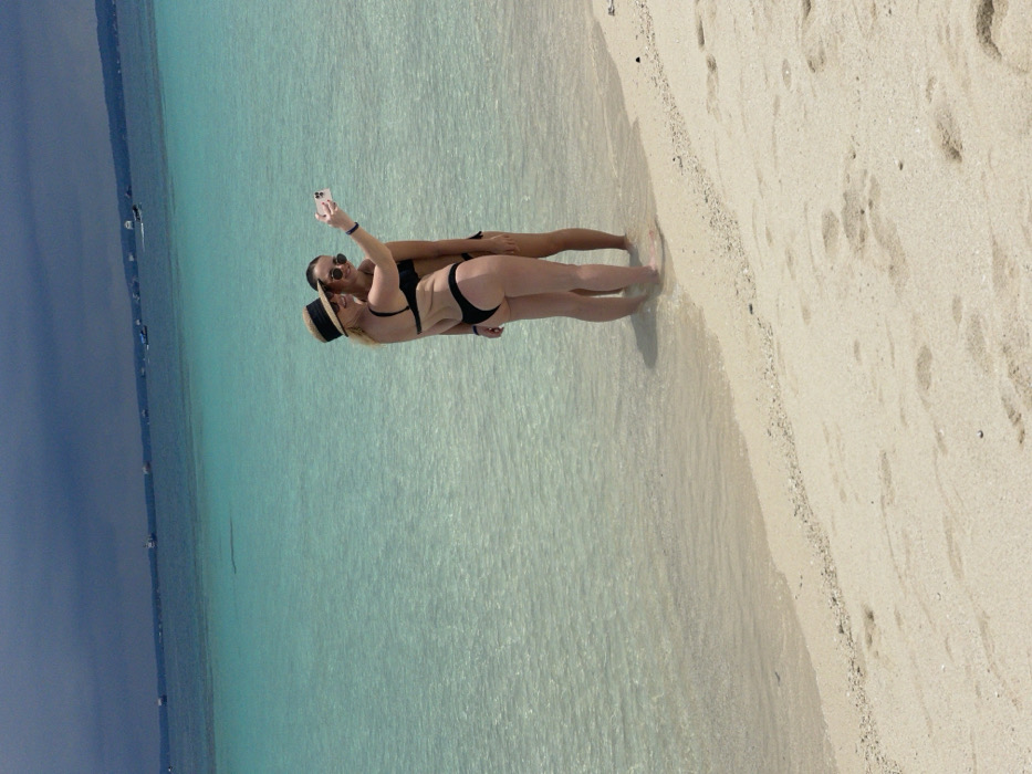 The image displays two women posing for a selfie on a sandy beach in Zanzibar, Tanzania. Both individuals wear black two-piece swimsuits and sunglasses. The woman positioned on the left, wearing a straw visor, extends her right arm to hold a light-colored smartphone, directed towards herself and the second woman. She stands with her lower calves submerged in clear, shallow turquoise ocean water. The second woman stands slightly behind her, also in the shallow water, facing the smartphone. The immediate foreground consists of light-colored sand with faint footprints. The background features a calm, transparent turquoise ocean extending to the horizon, dotted with multiple small boats. Above the distant ocean, the sky is dark and overcast.