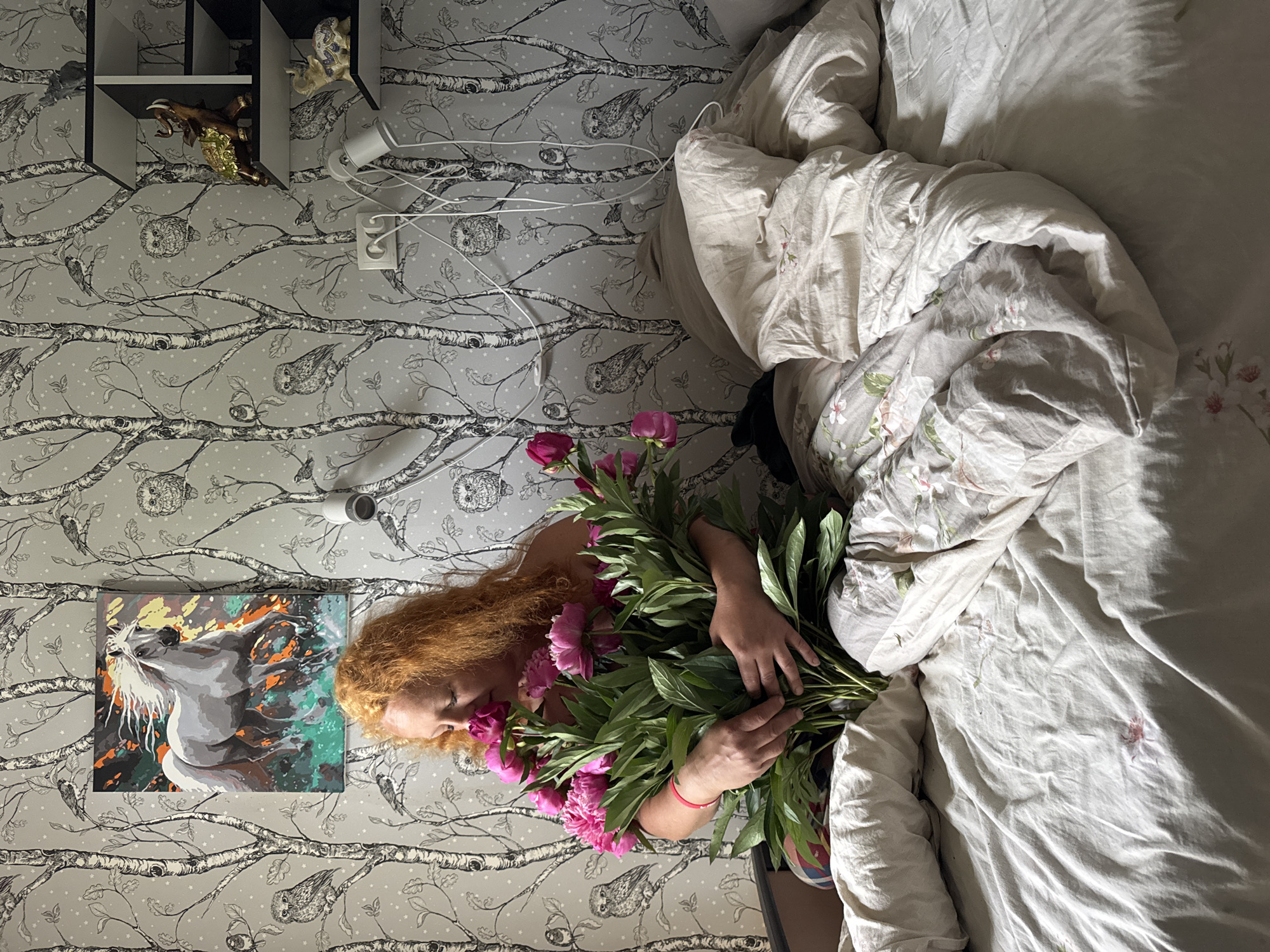 In a bedroom with wallpaper featuring a black and white birch tree and owl motif, a woman with vibrant red hair is seated on a bed. She is holding a large bouquet of vibrant pink peonies, with her face gently lowered towards them as if to smell their fragrance. The bed is covered with white and patterned bedding, and to the right, a shelf unit is visible on the wall, adorned with decorative elephant figurines. The overall scene suggests a moment of quiet enjoyment and appreciation of nature's beauty within a domestic setting. This room is located in a residential apartment in Chaikovsky, Russia.