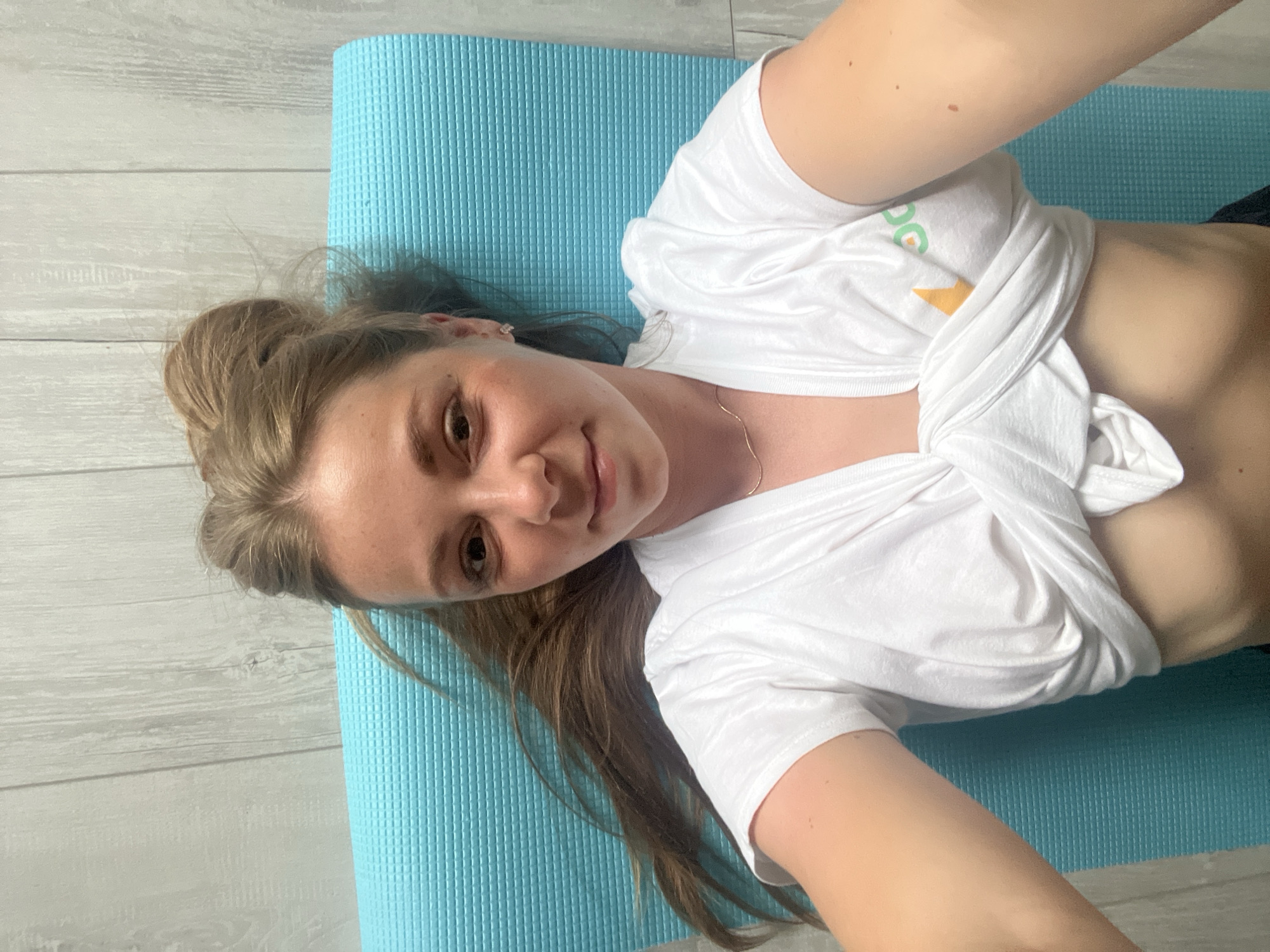 A young woman with blonde hair tied up in a bun lies on her back on a bright blue yoga mat. She is wearing a white t-shirt that is tied around her waist, exposing her toned stomach. Her hair is long and extends down her back, some of it resting on the mat. The yoga mat is placed on a light-colored wooden floor. The woman is looking directly at the camera with a slight smile. The lighting suggests it is daytime, and there are no indications of specific weather conditions. The image appears to be a selfie taken indoors. There is a subtle logo on her t-shirt, partially visible, with green and yellow elements. The overall mood is relaxed and informal.