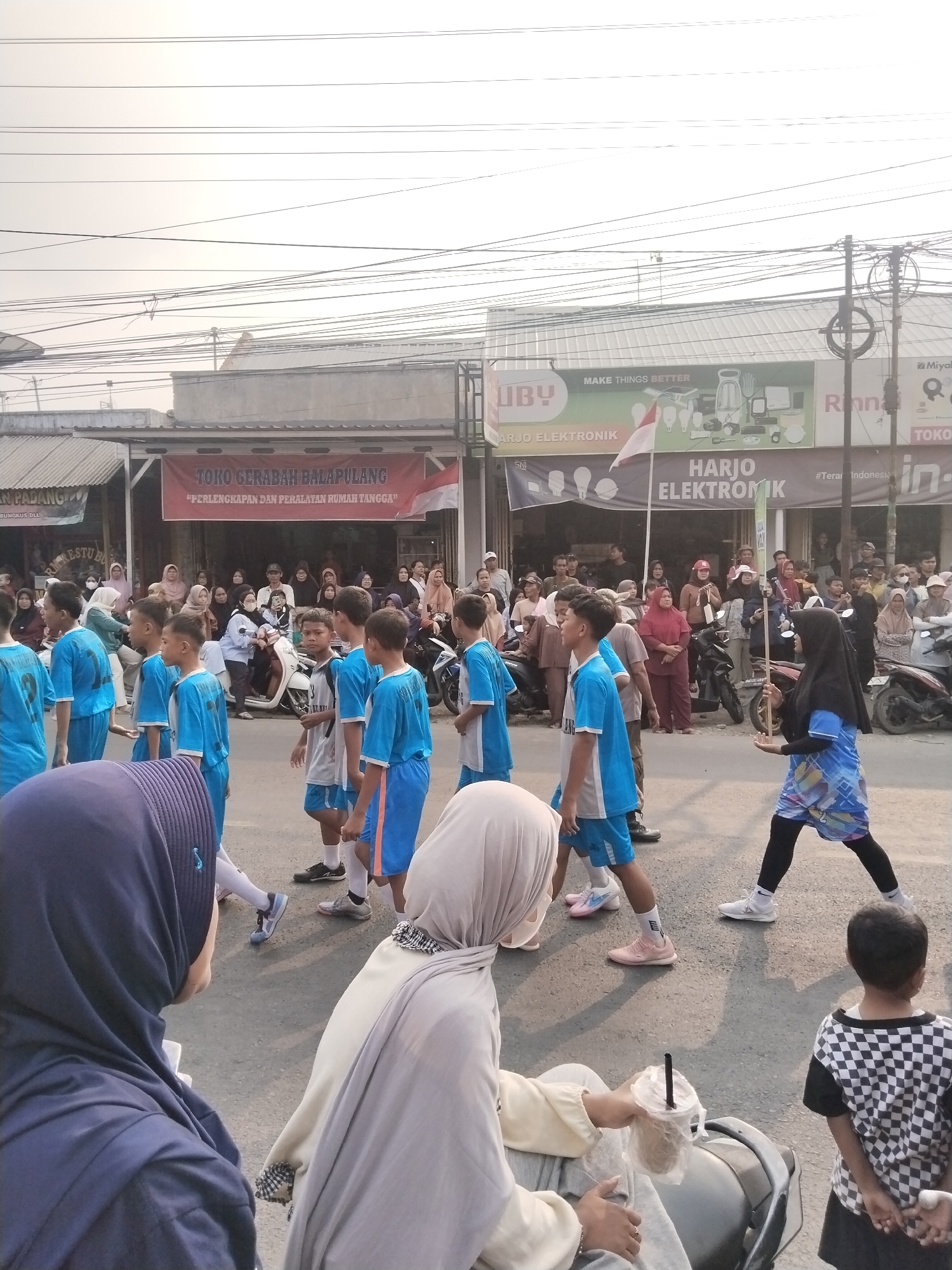 The image depicts a street scene in Balapulang Kulon, Indonesia, likely during a parade or event. A group of young boys in blue and white athletic wear are marching in the street, some looking towards the camera. They appear to be part of a school or sports team. In the background, several adults are gathered, watching the procession. Some are standing on the sidewalk, while others are seated or leaning against buildings. There are also several motorcycles parked along the street. The buildings in the background have signs advertising businesses, including "TOKO GERABAH BALAPULANG" and "HARJO ELEKTRONIK." The sky is overcast, and the overall lighting suggests it is daytime. The street is paved, and there are overhead power lines typical of an urban or semi-urban environment. One individual in the foreground, wearing a dark blue hijab, is partially visible from behind, holding a drink. Another individual in a light-colored hijab is also in the foreground.