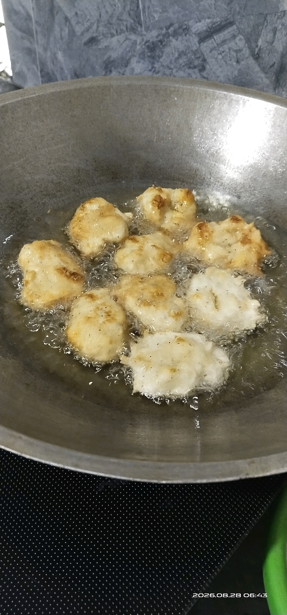 A close-up, overhead shot shows eight pieces of food being deep-fried in a large, round, metal wok filled with bubbling oil. The food items appear to be pale golden brown, suggesting they are either battered and fried, or a type of fritter. Some pieces have darker brown spots, indicating they are crisping up. The wok is set on a dark, textured surface, possibly a stovetop, which has a pattern of small white dots. The background is blurred, but appears to be a wall with a textured, gray pattern. The lighting is overhead and creates a sheen on the oil. The timestamp "2026.08.28 06:43" is visible in the bottom right corner of the image, indicating the date and time the photograph was taken. The scene is domestic and appears to be a kitchen environment. There is no information provided about the location in Matabu, Indonesia.