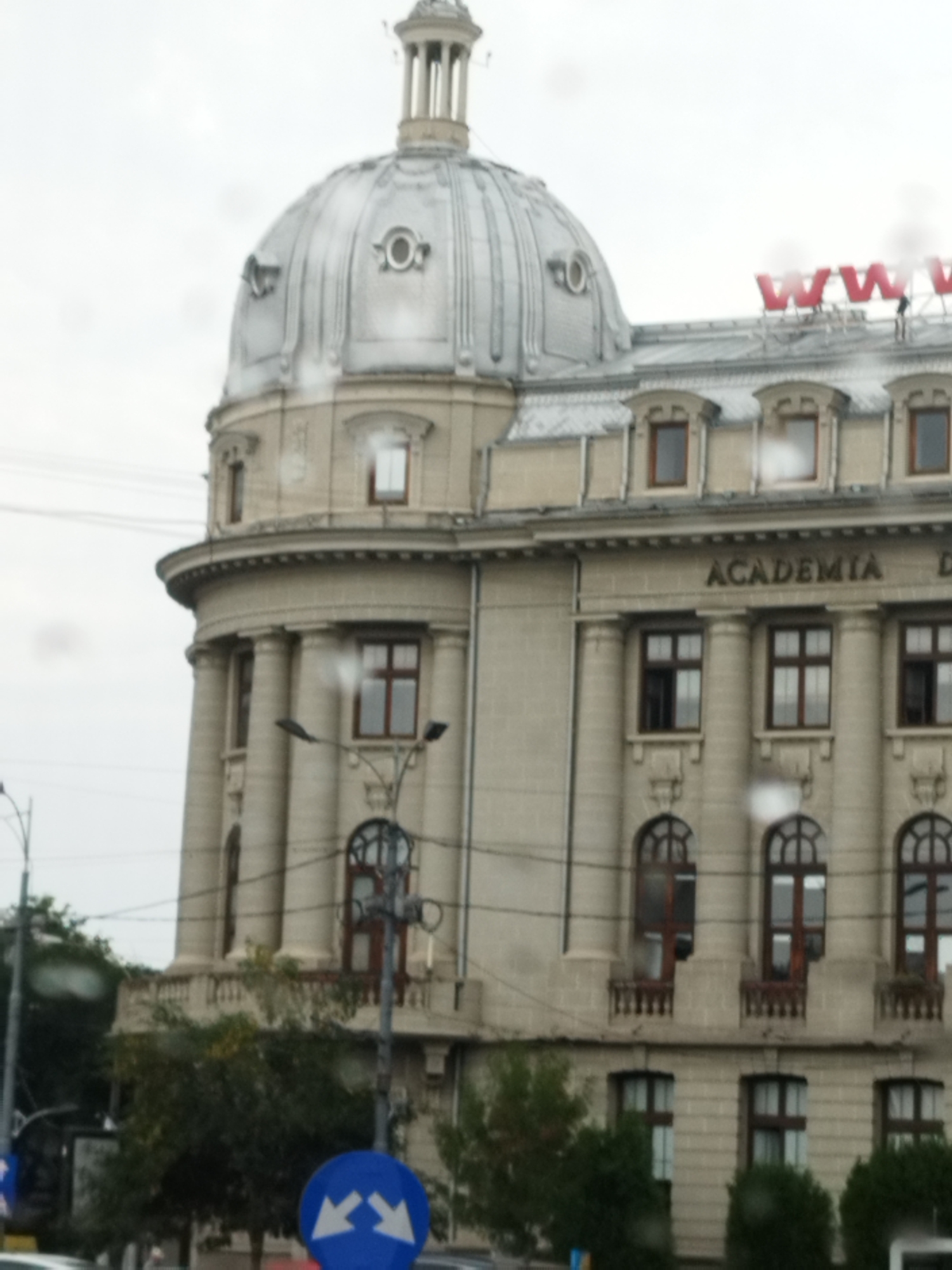 The image captures a portion of the Romanian Academy building in Bucharest, Romania, under a cloudy sky with visible raindrops on the lens. The grand architecture features a prominent dome, several windows with dark frames, and decorative columns. The word "ACADEMIA" is clearly visible inscribed on the building's facade. Red lettering, possibly a sign, is present on the roof. The lower part of the image shows trees, streetlights, and traffic signs, indicating an urban setting. One sign is a blue circular sign with two white arrows pointing outwards, suggesting a traffic direction or lane change instruction. The overall scene depicts a landmark building in a city environment on a potentially wet day.