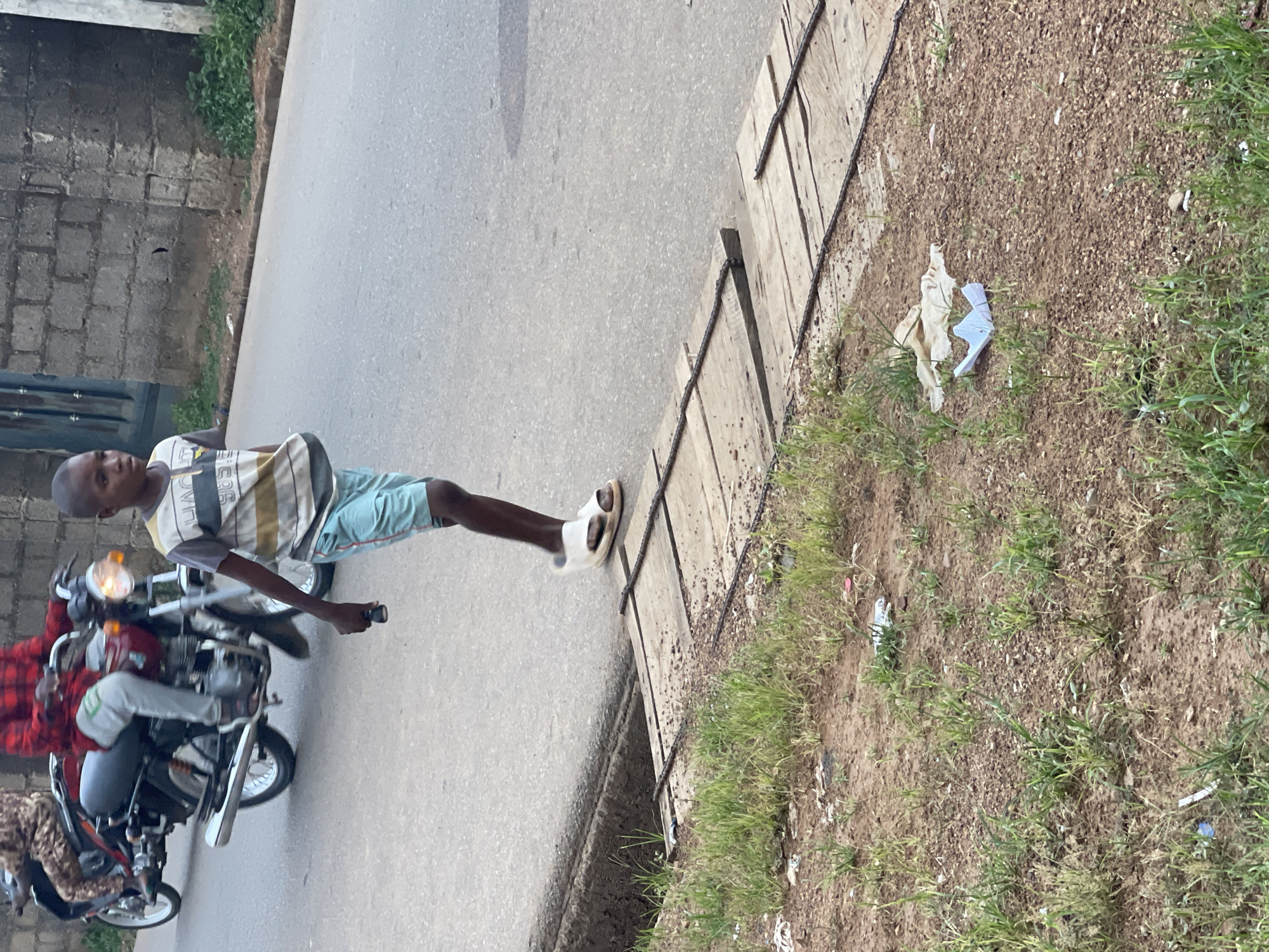 In Akwanga, Nigeria, a young man is seen standing on the roadside next to a motorcycle. He is wearing a striped shirt, blue shorts, and sandals. The motorcycle is red and has two people on it. The road is asphalt, and there is a concrete curb with wooden planks alongside. The ground adjacent to the curb is covered in dirt, grass, and trash. A brick wall is visible in the background.