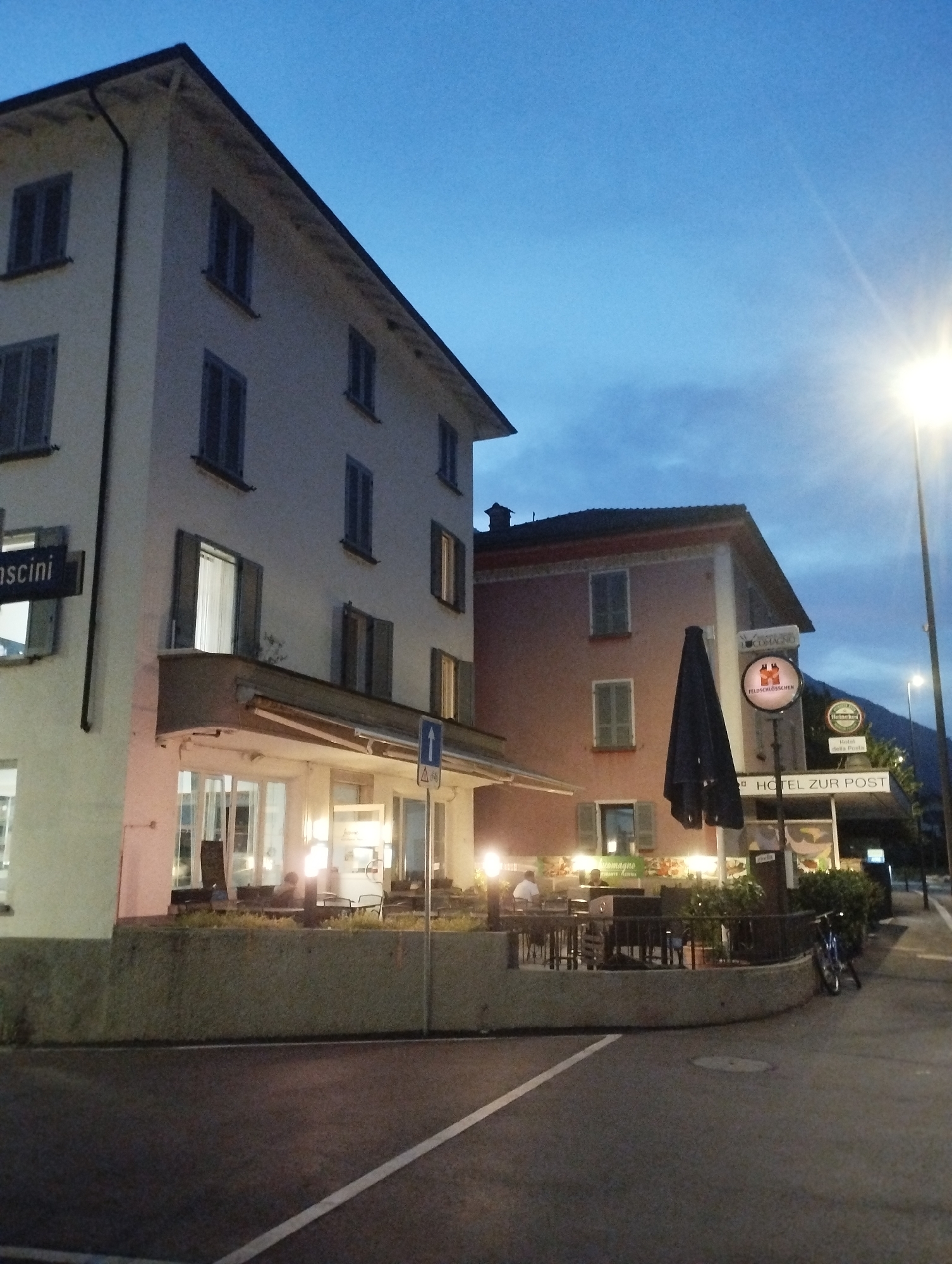 The image depicts a street scene in the evening or dusk, with artificial lights illuminating the buildings and outdoor seating areas. A two-story building with white and pink facades is prominent, featuring multiple windows with closed shutters. The ground floor of the white building has a covered outdoor seating area with tables and chairs, suggesting a restaurant or cafe. Some patrons are visible sitting at the tables. A sign with "VASCINI" is visible on the white building.
Adjacent to the white building is a pink building, also with multiple windows. This building has a sign that reads "HOTEL ZUR POST" and other smaller signs, including one for "Heineken". A large, dark umbrella is positioned in front of the pink building, partially obscuring some of the signage.
In the foreground, a paved area with white lane markings suggests a road or sidewalk. A bicycle is parked to the right of the pink building. The sky is a deep blue with some clouds, and a bright street lamp is visible on the far right, casting a strong light. The overall atmosphere is tranquil and suggestive of a European town or village.