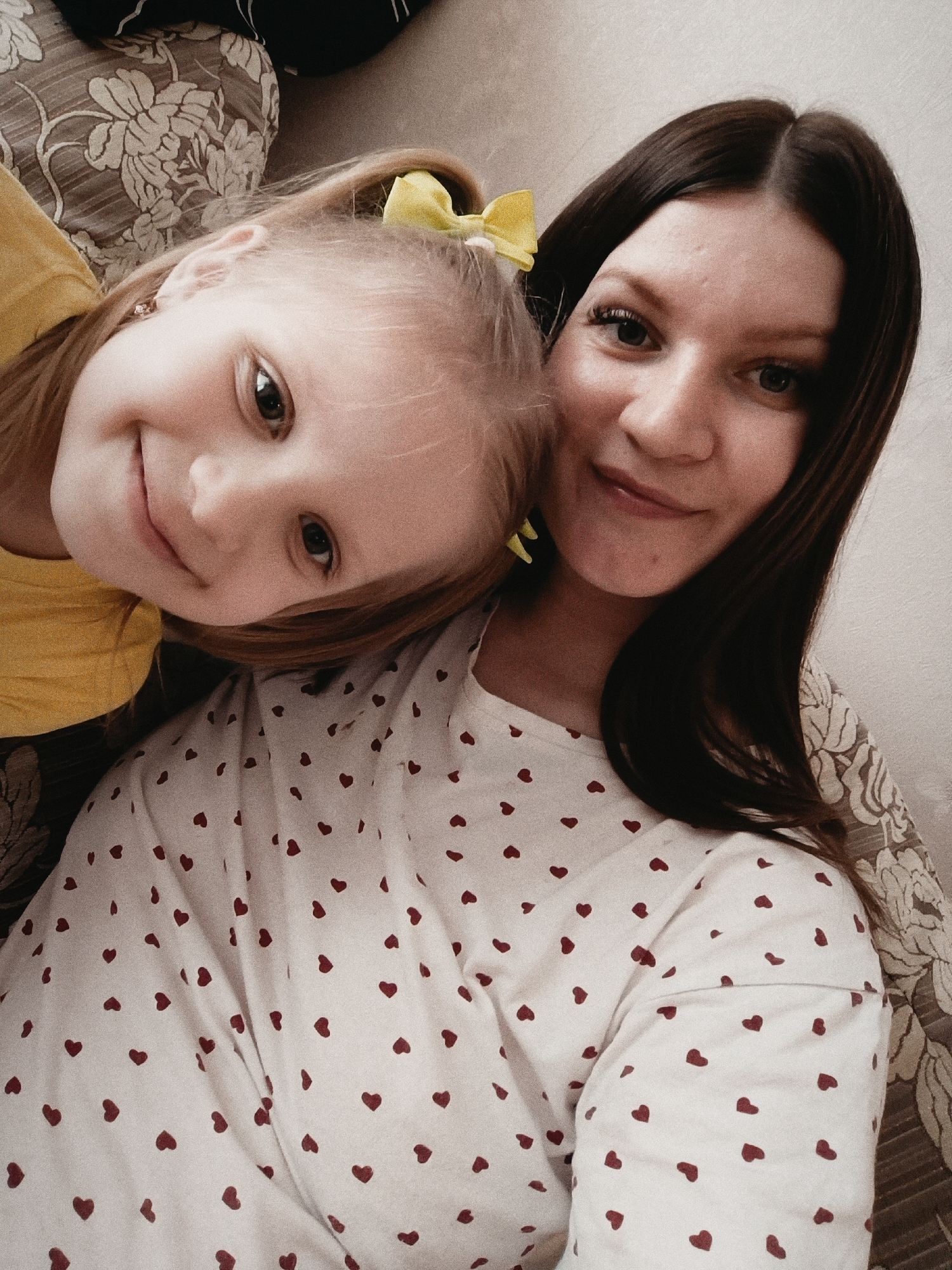 The image features an adult female and a young female child positioned closely together in an indoor setting. The adult female, located on the right, has long dark brown hair with a middle part, dark eyes, and a subtle smile while looking directly at the camera. She wears a white long-sleeved top patterned with numerous small red hearts. The young female child, positioned on the left and partially overlapping the adult, has light-colored hair secured with a yellow bow and a small earring visible in her right ear. She is wearing a yellow top and also smiles directly at the camera, resting her head against the adult. The background consists of a plain light-colored wall or surface on the left and a patterned fabric, possibly upholstery, with a floral motif in muted tones on the right. This scene is identified as being located in the city of Tobolsk, Russia.