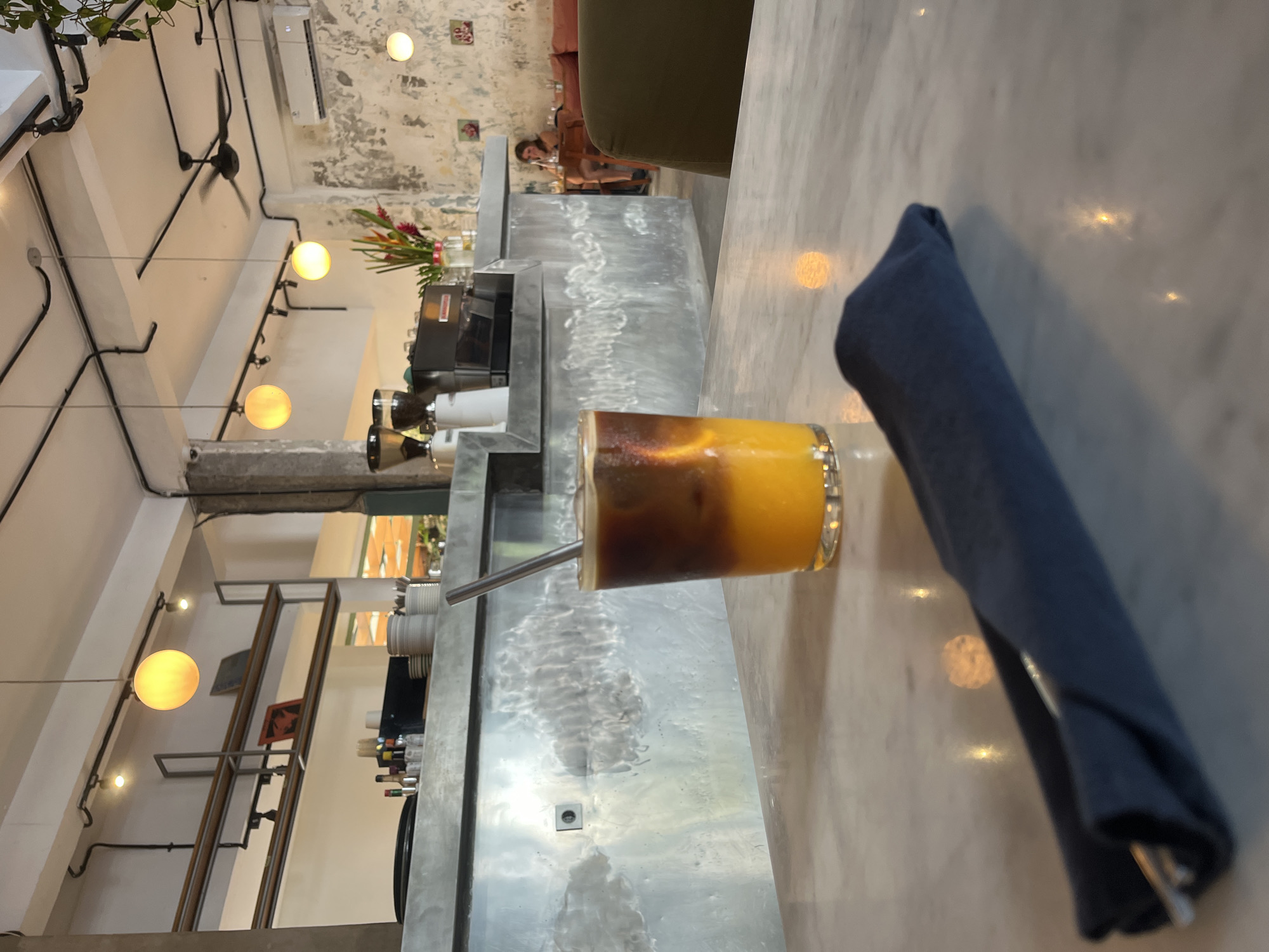 The photograph captures a scene inside a cafe or restaurant, potentially located in North Kuta, Indonesia. The main subject is a glass of liquid, possibly a coffee drink, with distinct layers of dark and light-colored liquids. A metal straw is inserted into the glass. To the right of the glass is a folded dark blue napkin. The drink and napkin sit on a marble-like countertop.
In the background, behind the counter, there are coffee-making machines, bottles, and flowers arranged in a vase. A person is sitting at a table further back in the room. The interior design features exposed pipes, hanging light fixtures, and shelving units. The lighting is a mix of natural and artificial light. The walls are textured.