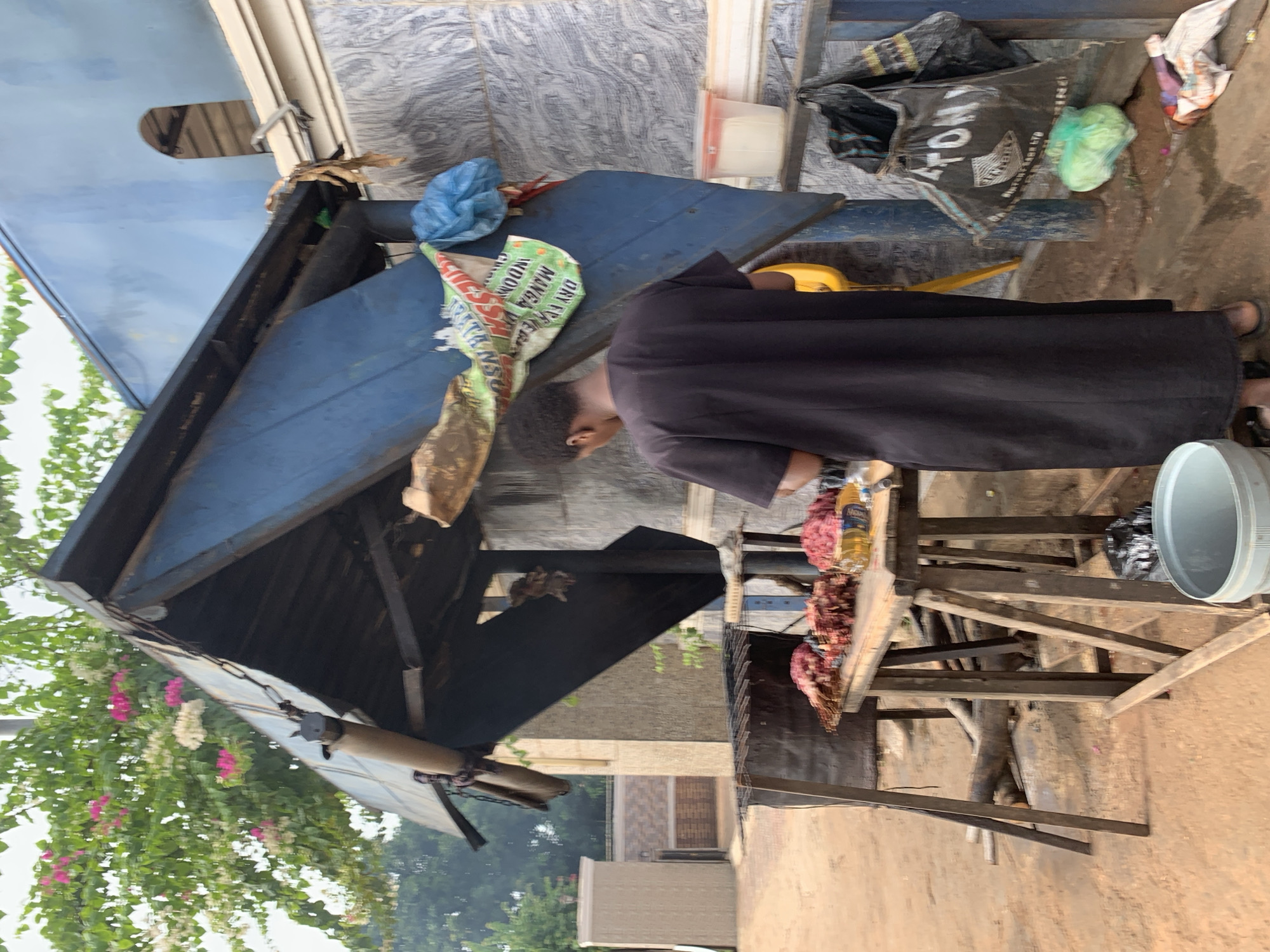 A person wearing a black robe stands next to a grill in what appears to be an outdoor setting in Nsugbe, Nigeria. The grill, constructed of wood and metal, is laden with raw meat. The person is looking down towards the grill. A blue and black makeshift shelter is above them. Next to the grill is a white bucket. A bag of animal feed, blue plastic bag, and other debris are in the background. Trees are visible in the distance.