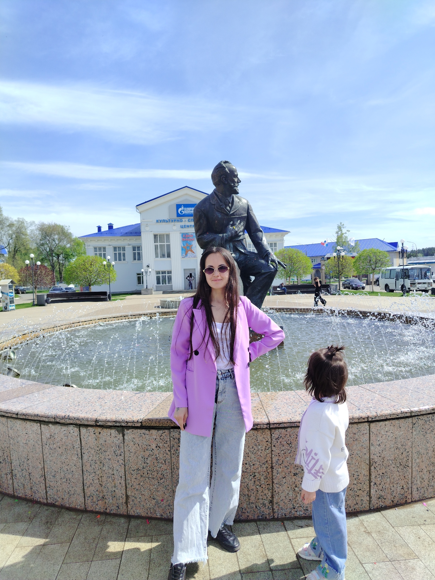 The photograph captures a scene in the city of Chaikovsky, Russia, in front of the Cultural Center (Kультурно-Спортивный Центр) of Gazprom Dobycha (Газпром добыча) company, identifiable by the company logo on the building’s facade. A bronze statue of a seated man is prominently positioned in the foreground. In front of the statue, a woman wearing a lilac blazer, white top, and light-wash jeans stands with her hand on her hip and sunglasses on. To her right, a young girl with dark hair in a ponytail stands facing the fountain, wearing a white sweater and jeans. The setting includes a circular fountain in the center with water jets spraying upwards. The background also features trees, streetlights, benches, and a parked bus. The sky is partially cloudy.