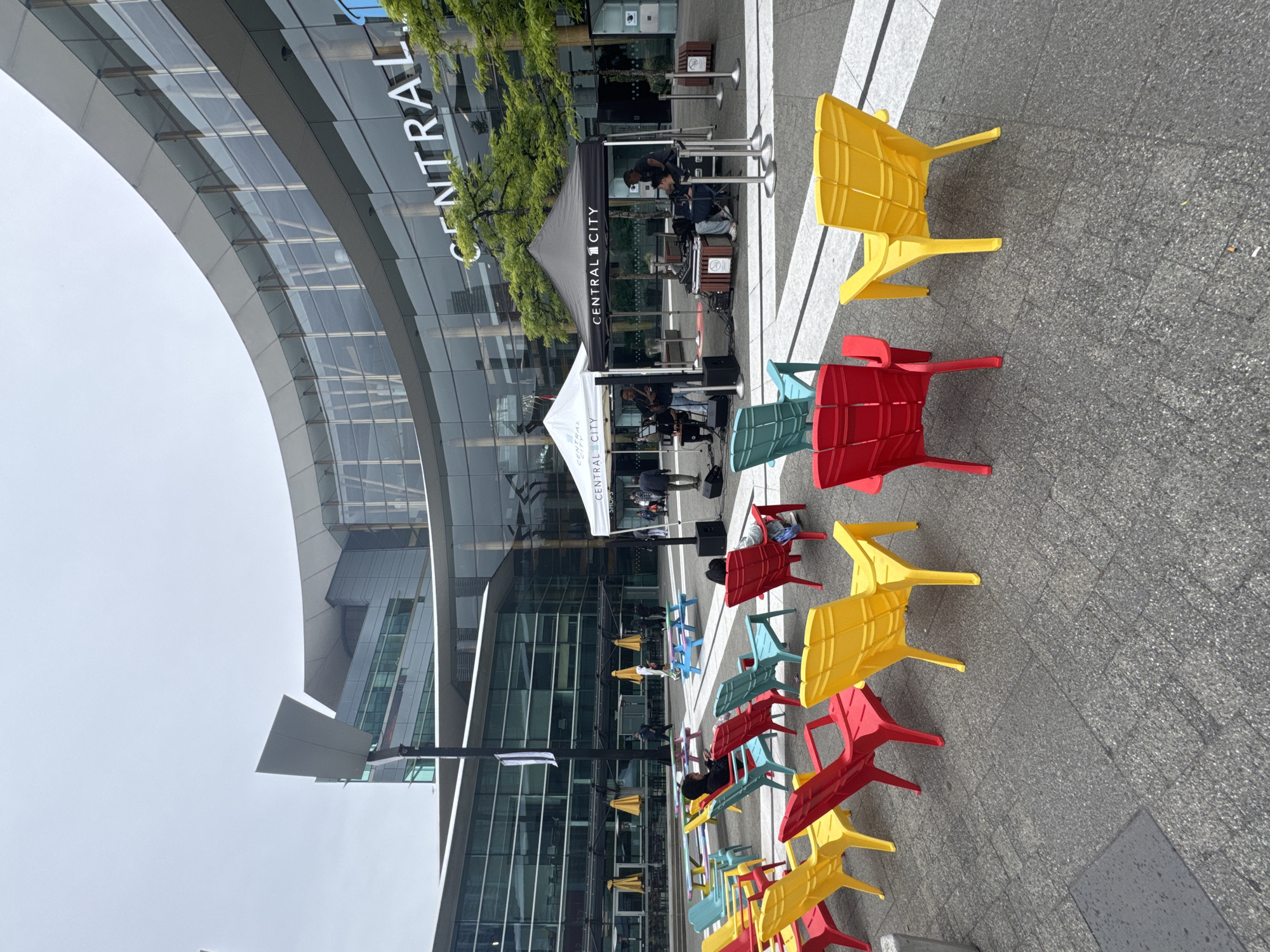 This image depicts an outdoor plaza in front of a modern building with a large, curved glass facade. The building has the prominent white lettering "CENTRAL CITY" visible on its exterior. The plaza is paved with large square tiles and features numerous brightly colored Adirondack-style chairs, predominantly in red, yellow, and teal.
In the mid-ground, two pop-up tents are set up. The tent on the left is white with "CENTRAL CITY" branding, and the tent on the right is black, also branded with "CENTRAL CITY." A live band, consisting of at least three individuals, is performing under the black tent. They are set up with musical equipment including amplifiers and a drum kit. Several people are seated in the colorful chairs, observing the performance or relaxing. Others are walking in the background.
Scattered throughout the plaza are picnic tables, some with umbrellas providing shade. The overall atmosphere suggests a public gathering space or event. The environment is overcast, with a muted sky. The location is identified as Central City in Surrey, Canada, a well-known shopping center and office complex.