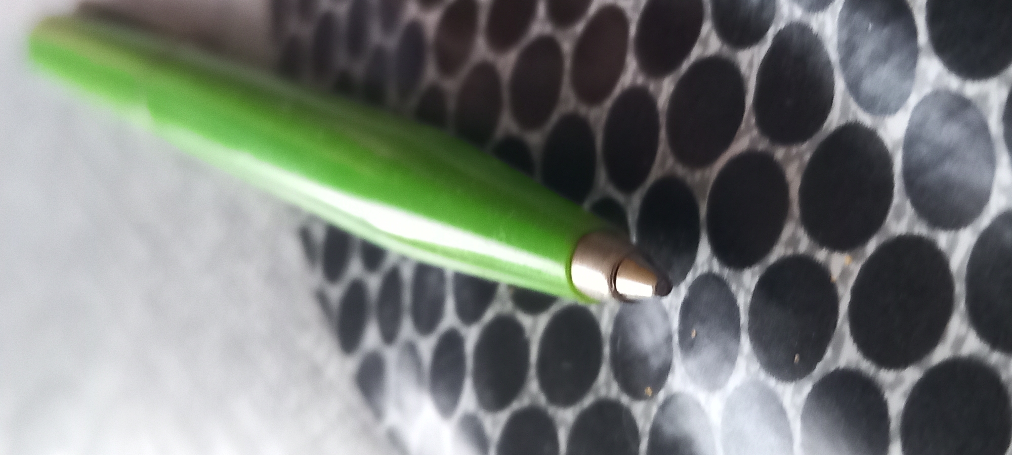 A vibrant green ballpoint pen rests diagonally on a surface patterned with numerous dark, ovular shapes against a lighter, possibly white or light grey, background. The metallic tip of the pen, appearing bronze or gold, is in sharp focus, showcasing the dark writing point. The pen body is a bright, solid green, reflecting some light along its length. The patterned surface extends across the lower and middle portions of the frame, with the upper portion of the image exhibiting a heavily blurred, light grey background. The image's shallow depth of field sharply isolates the pen tip and the immediate surrounding pattern from the indistinct background elements. No individuals, specific actions, or interactions are discernible. The environment appears to be an indoor setting; however, the image provides no visual information to determine its geographical location, including the city of Kryvyi Rih, Ukraine.