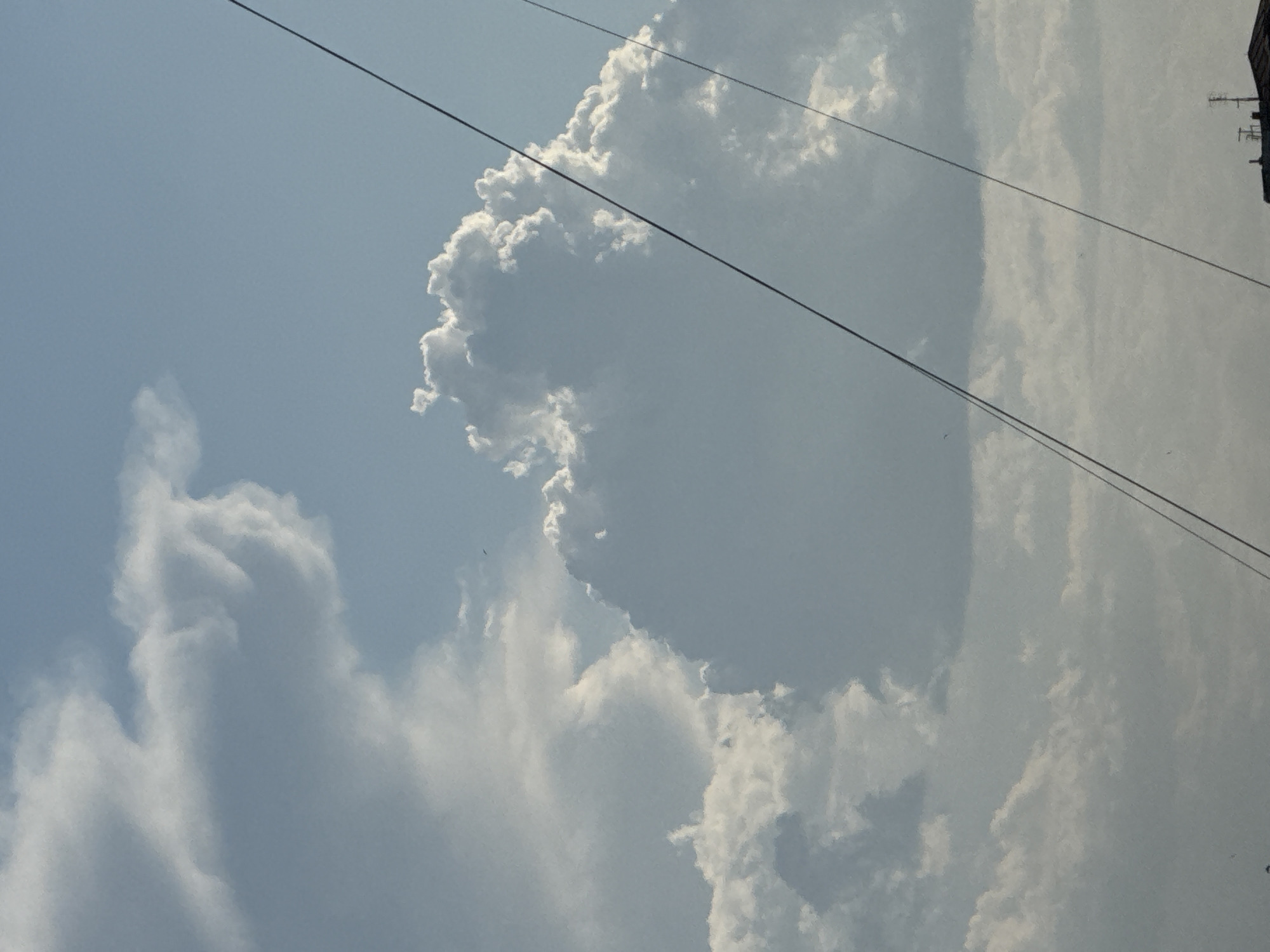 A low-angle, vertical shot captures a sky filled with clouds and two dark, thin lines that appear to be wires. The clouds are mostly white and puffy, with some areas of shadow indicating depth. The sky is a light blue. In the bottom right corner, a sliver of a dark roof with antennas is visible, suggesting an urban or suburban environment. The overall scene is serene and observational. The image is taken from the city of Abovyan, Armenia, looking upwards at the sky.